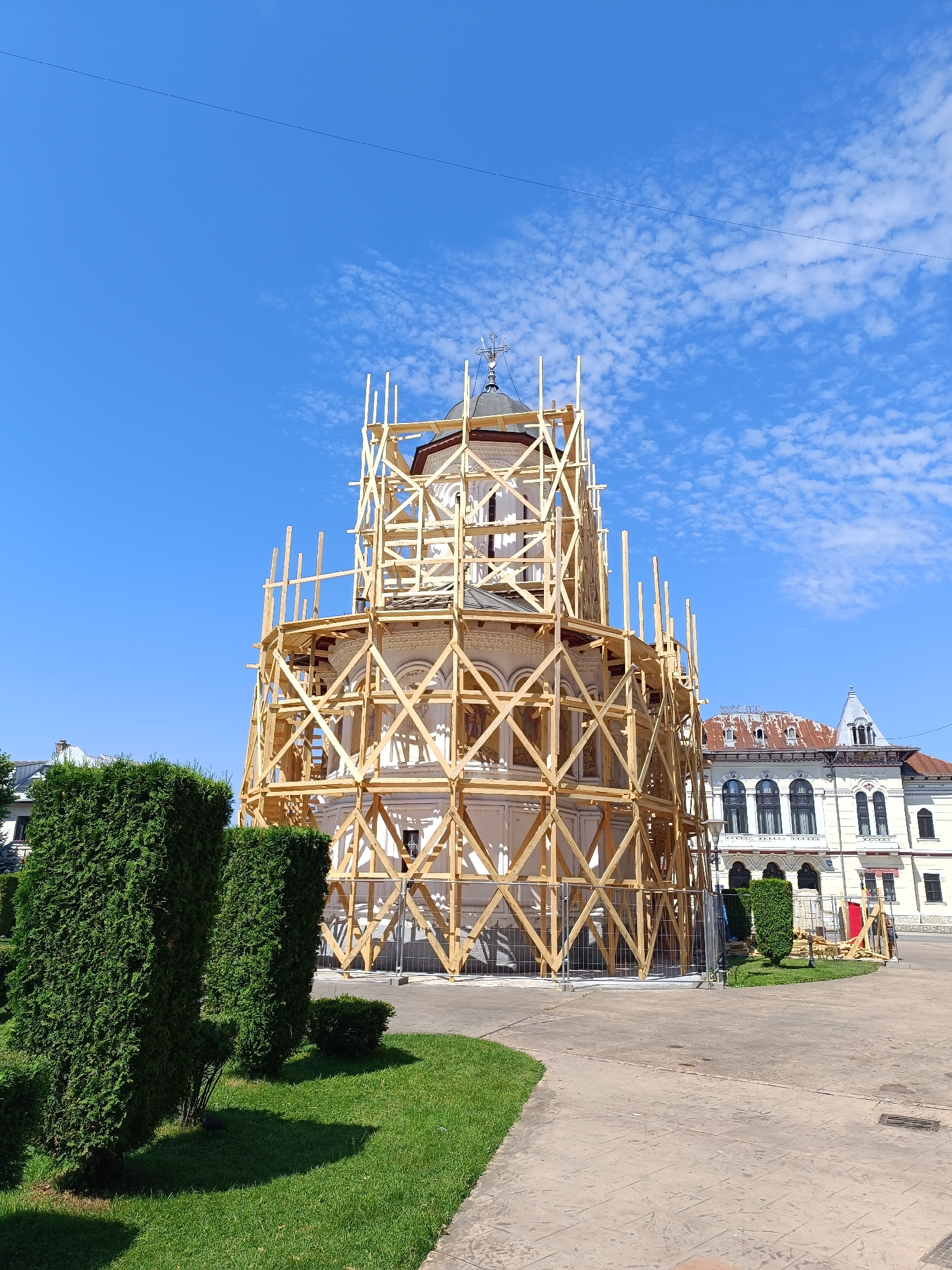 The image shows the Clock Tower of Saint John the Baptist in Târgu Jiu, Romania, undergoing restoration. The tower, a prominent architectural landmark, is encased in extensive wooden scaffolding, suggesting ongoing repair or maintenance work. The scaffolding covers most of the tower's facade, obscuring its details but revealing its cylindrical shape. Above the scaffolding, the top of the tower, including its dome and cross, is visible.
The tower is situated in an open, paved area, likely a public square. In the foreground, manicured green lawns and tall, sculpted evergreen bushes are visible, indicating a well-maintained park or garden area surrounding the historical site. Behind the tower and to its right, other buildings are present. One building, possibly a historical structure itself, features arched windows and decorative architectural elements, with a weathered, reddish-brown roof. The sky above is a clear, bright blue with scattered white clouds. No people are visible in the image. The scene appears to be a sunny day, indicative of outdoor public space in a Romanian city.