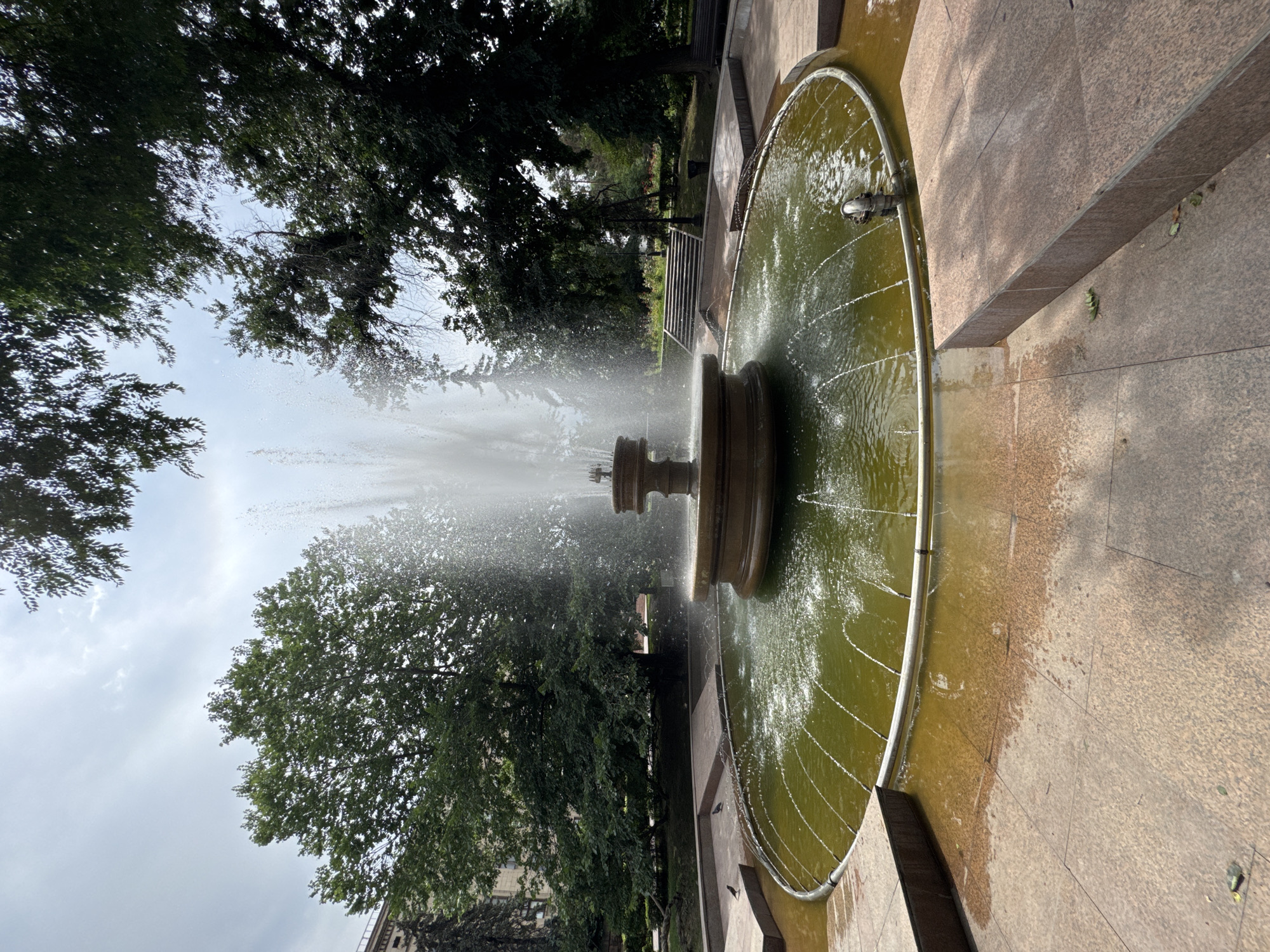 An active, multi-tiered fountain with a central vertical spray is situated in a park setting. The fountain's circular basin contains murky green water, with multiple smaller jets creating a ring of water sprays at the basin's edge. The fountain is surrounded by a paved area with large, square granite tiles and raised rectangular platforms. Beyond the fountain, lush green trees create a canopy overhead, partially obscuring a cloudy sky. In the background, a large building with a light-colored facade is visible, and a set of concrete stairs leads up a slight incline into the park. The overall scene depicts a public park area with a prominent water feature. This is likely located in the central Almaty region of Kazakhstan due to the architectural style of the building and the commonality of such park features in urban environments there.