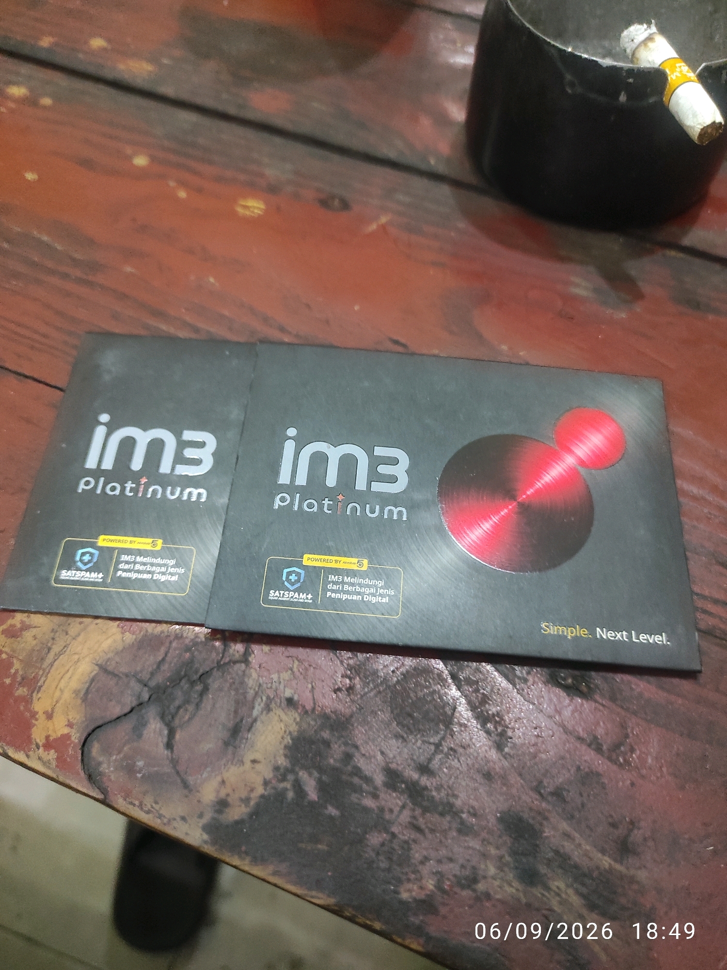 The image shows two dark grey cards with the text "im3 platinum" printed on them. The cards are lying on a weathered wooden surface that appears to be a table. In the background, an ashtray with a lit cigarette is visible. The lighting in the image suggests it was taken indoors. The bottom right corner of the image shows a timestamp: "06/09/2026 18:49".