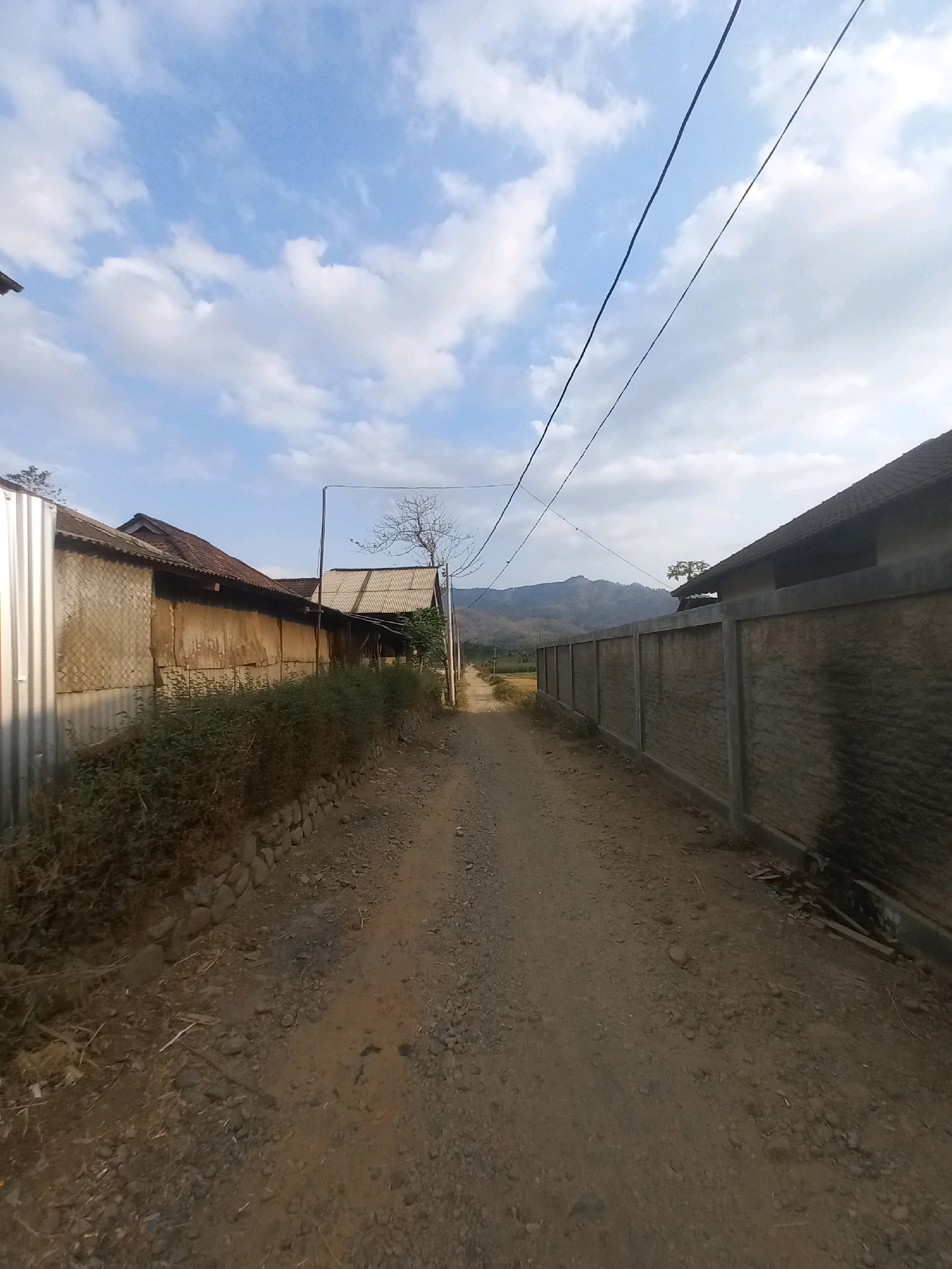 The image depicts a dirt road in the village of Girirejo, Indonesia. On the left side of the road, there are several structures with corrugated metal and woven material walls, and a low stone wall with dense green bushes growing behind it. On the right side of the road, a tall concrete wall runs along the property line. Overhead, power lines stretch across the sky, which is partially filled with white clouds against a blue background. In the distance, a mountainous landscape is visible. The road is unpaved and appears to be a path through a rural area. There are no people or animals visible in the scene. The overall impression is one of a quiet, somewhat rustic village environment.