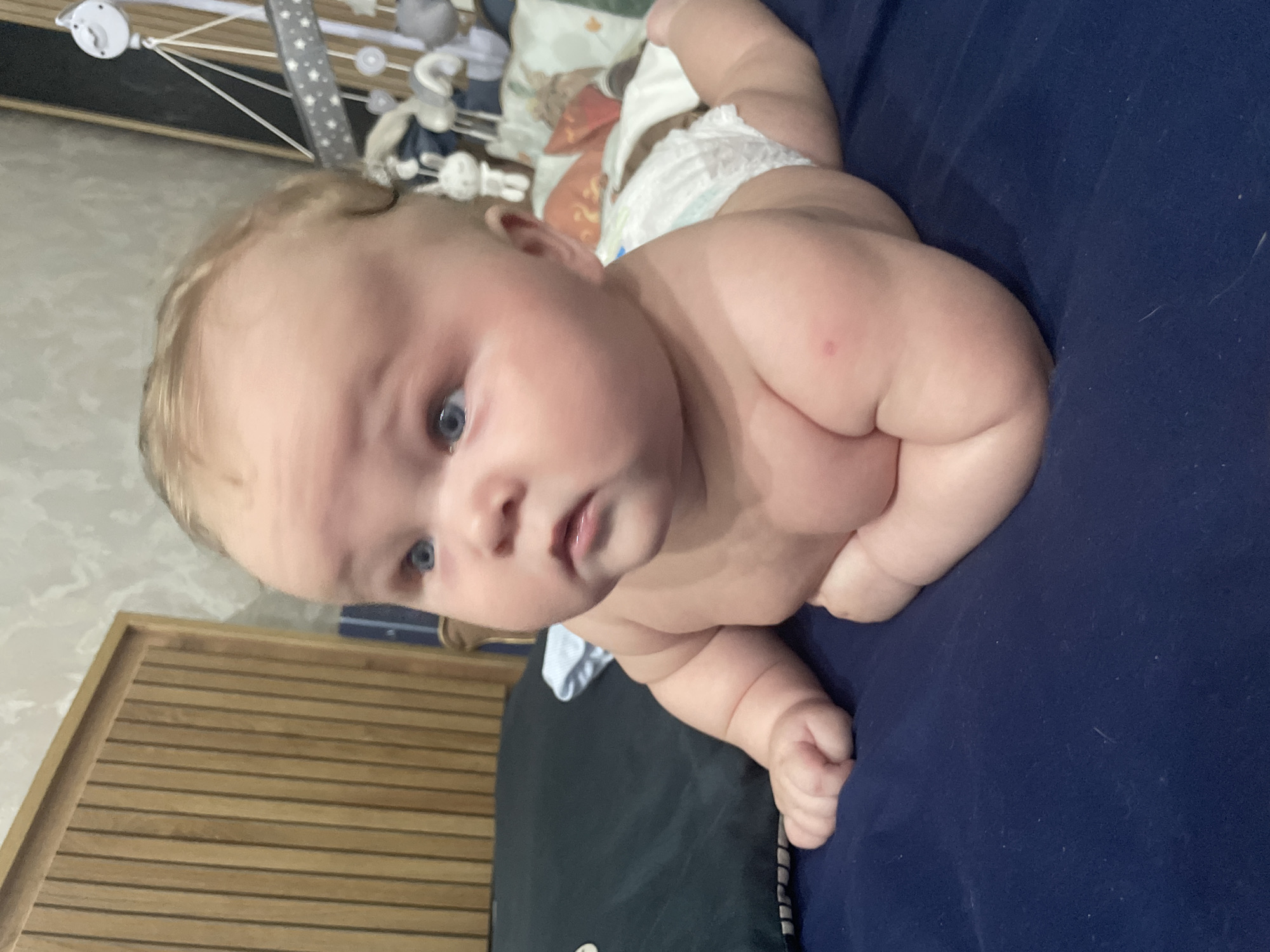 An infant, likely under one year old, is positioned on its stomach on a dark blue surface, possibly a bed or mat. The infant has fair hair, blue eyes, and is wearing a diaper. Its body is turned to the right, with its head also facing right and slightly upward, looking towards something out of frame. The background includes a wooden headboard with vertical slats, a wall with textured wallpaper, and a baby mobile with stuffed animals. There is also a dark pillow to the left of the infant and some bedding with a patterned pillow visible behind it. There is a small red mark on the infant's left shoulder. The image appears to be taken indoors. It is not possible to determine the location in Stavropol, Russia from the image provided.