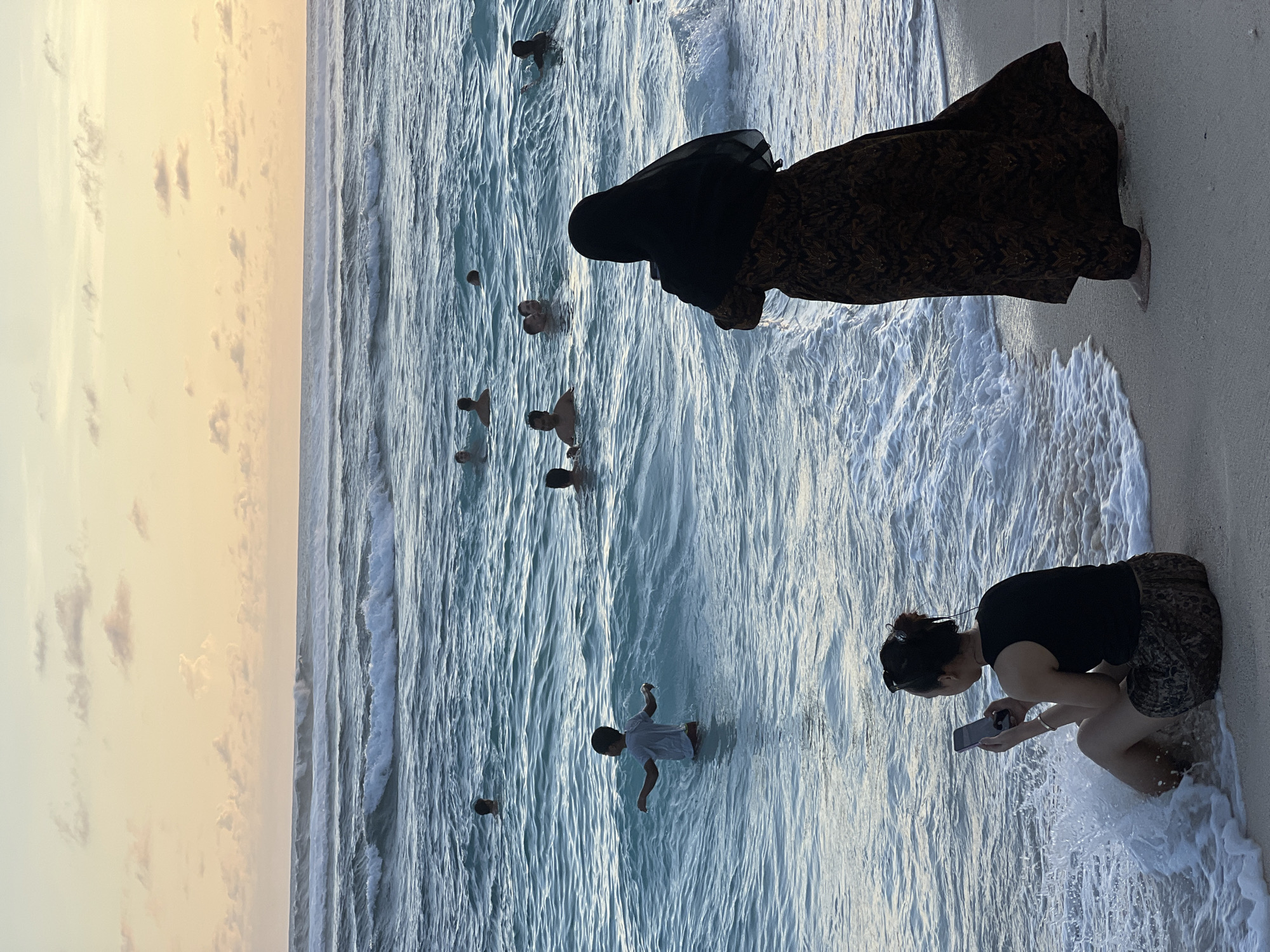 The image depicts a beach scene in Ungasan, Indonesia. The sky is a pale yellow with scattered clouds, transitioning to a blue ocean with visible waves. Several people are in the water, some swimming and others standing. On the shoreline, a woman in a black hijab and patterned dress is walking towards the water. Another woman is kneeling near the water's edge, looking at a mobile phone, with waves washing around her. The beach sand is light in color.