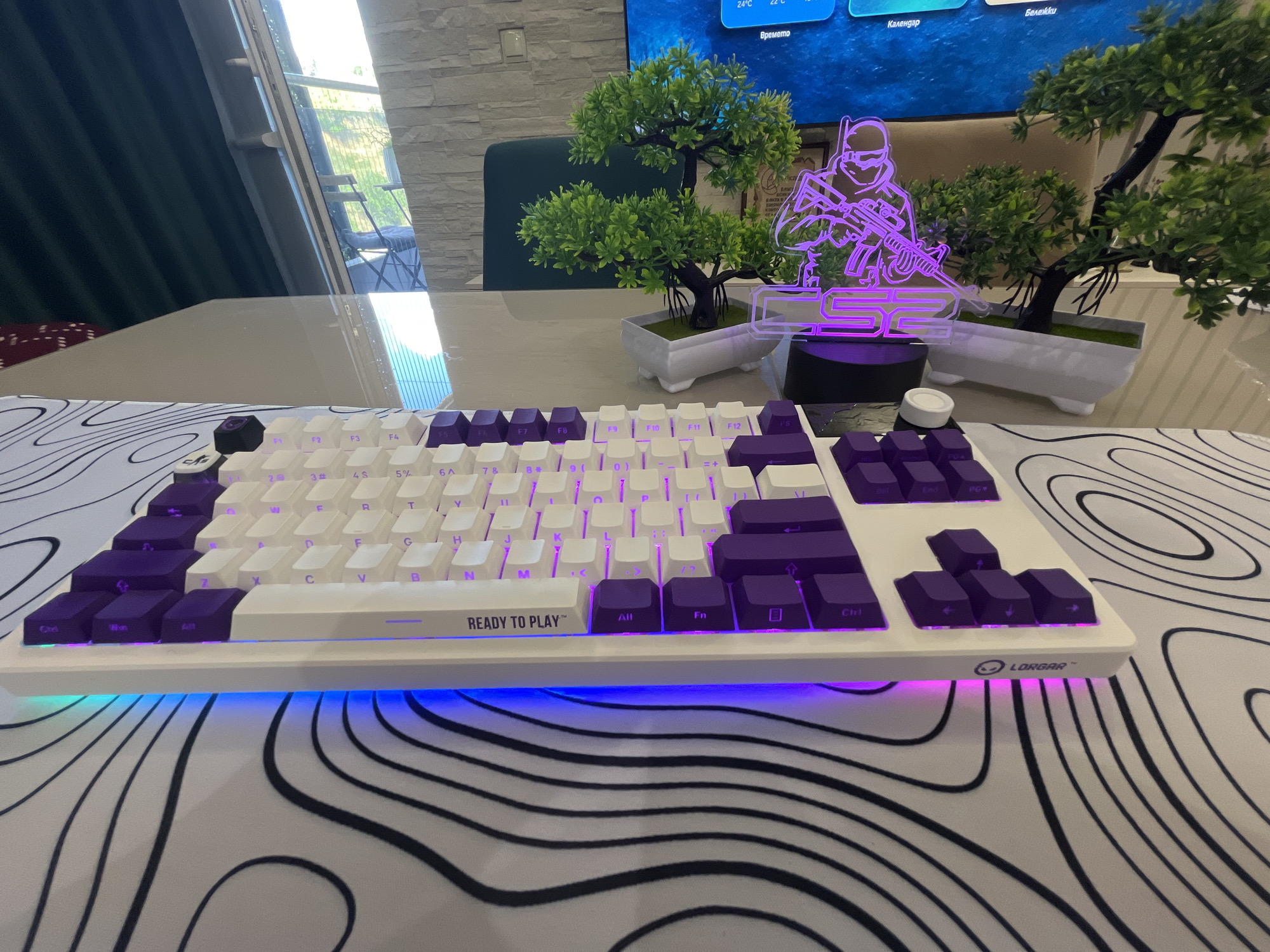 The image shows a white keyboard with purple and white keys, illuminated with purple and blue LED lights. The keyboard is placed on a desk with a black and white abstract pattern. In the background, there are two small artificial trees in white pots, and a neon sign depicting a character with a gun. A large screen displays weather information and other graphics. The scene appears to be an indoor setting, possibly a gaming or office setup, with a window showing greenery and furniture outside. The overall impression is of a modern, tech-oriented environment. There is no visible indication that this scene is located in Varna, Bulgaria.