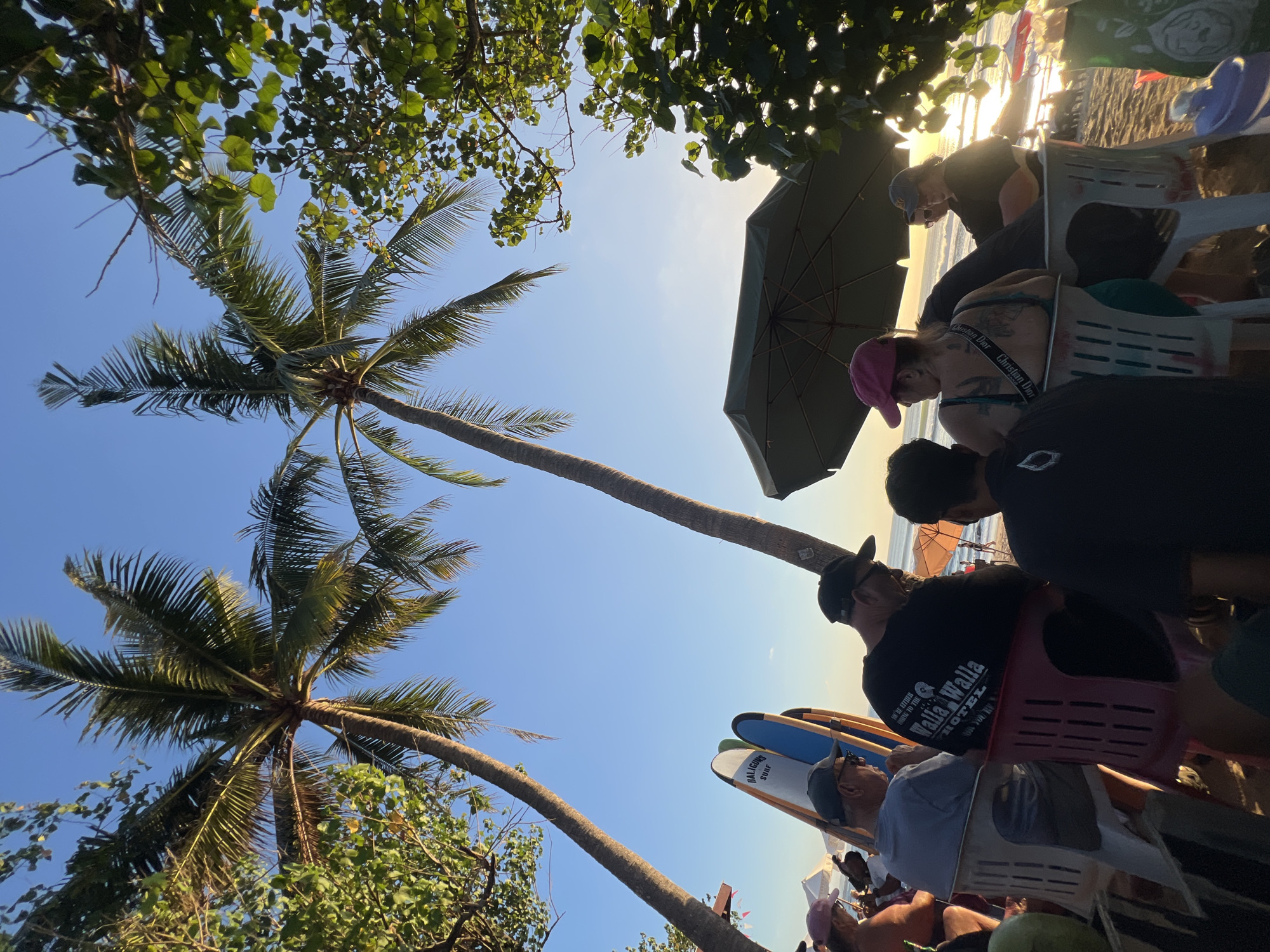 At a beach in Kuta, Indonesia, a group of people are seated on chairs under palm trees, facing the ocean during sunset. Two palm trees frame the scene, with their fronds reaching towards the clear blue sky. In the foreground, several individuals are visible, some wearing hats and casual attire. Surfboards are propped up behind them, suggesting a surfing environment. A large umbrella provides shade for some of the seated individuals. The sun is low on the horizon, casting a warm glow across the beach and water, with gentle waves visible in the distance. Other beachgoers and umbrellas can be seen further down the shoreline.