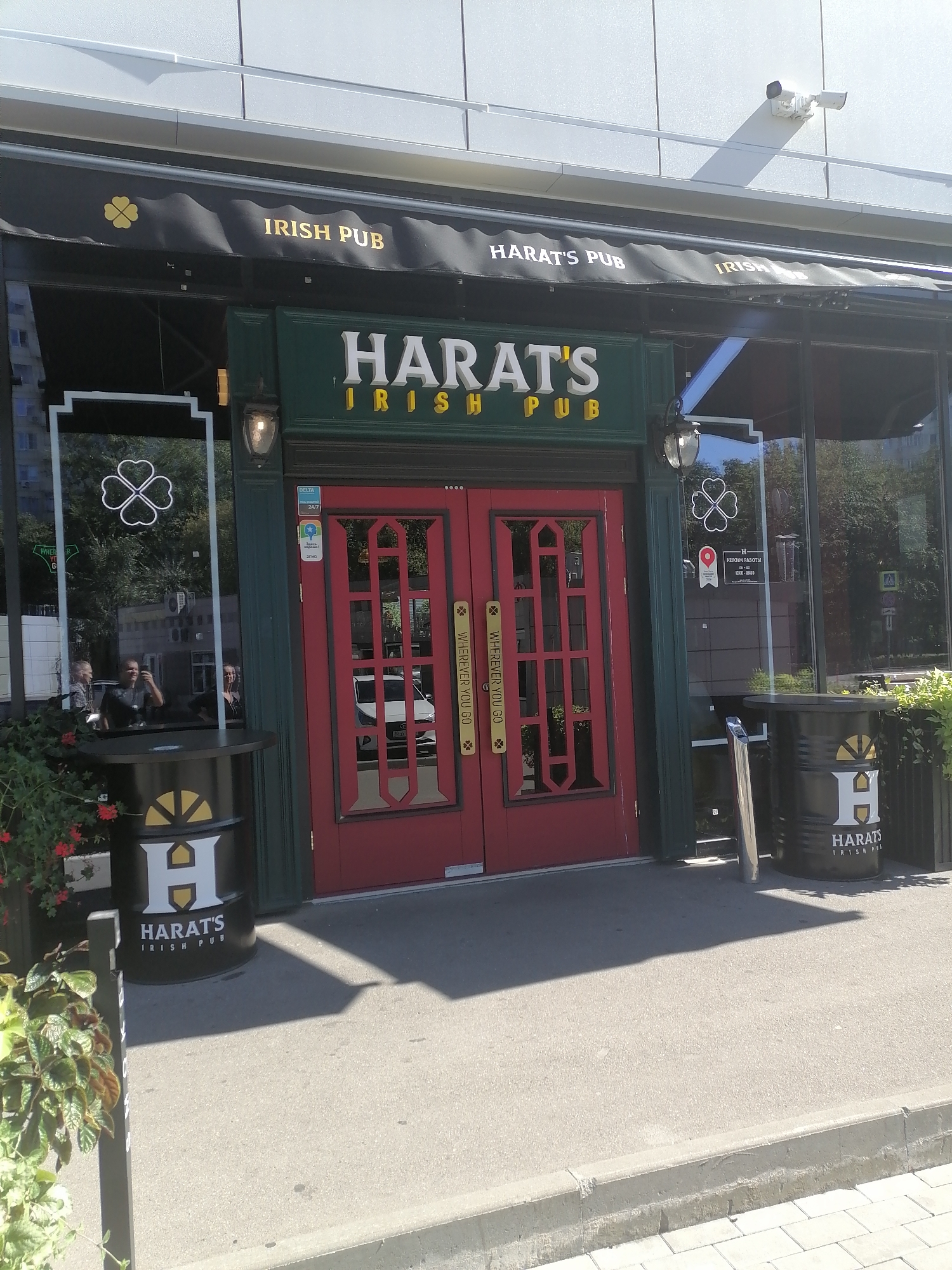 The image shows the entrance to a "Harat's Irish Pub" in Moscow, Russia. The facade is dark green with red double doors and gold lettering. Above the entrance, an awning displays "IRISH PUB" and "HARAT'S PUB" in gold. Two large, black cylindrical trash receptacles with the "Harat's Irish Pub" logo are placed on either side of the entrance. One of the trash receptacles has a prominent white "H" logo on it, with gold accents and the words "HARAT'S IRISH PUB" below. A security camera is visible mounted on the wall above the awning. Reflections in the glass windows show greenery and some building structures. The ground in front of the pub is paved with concrete and there is a curb with paving stones. The lighting suggests it is daytime.