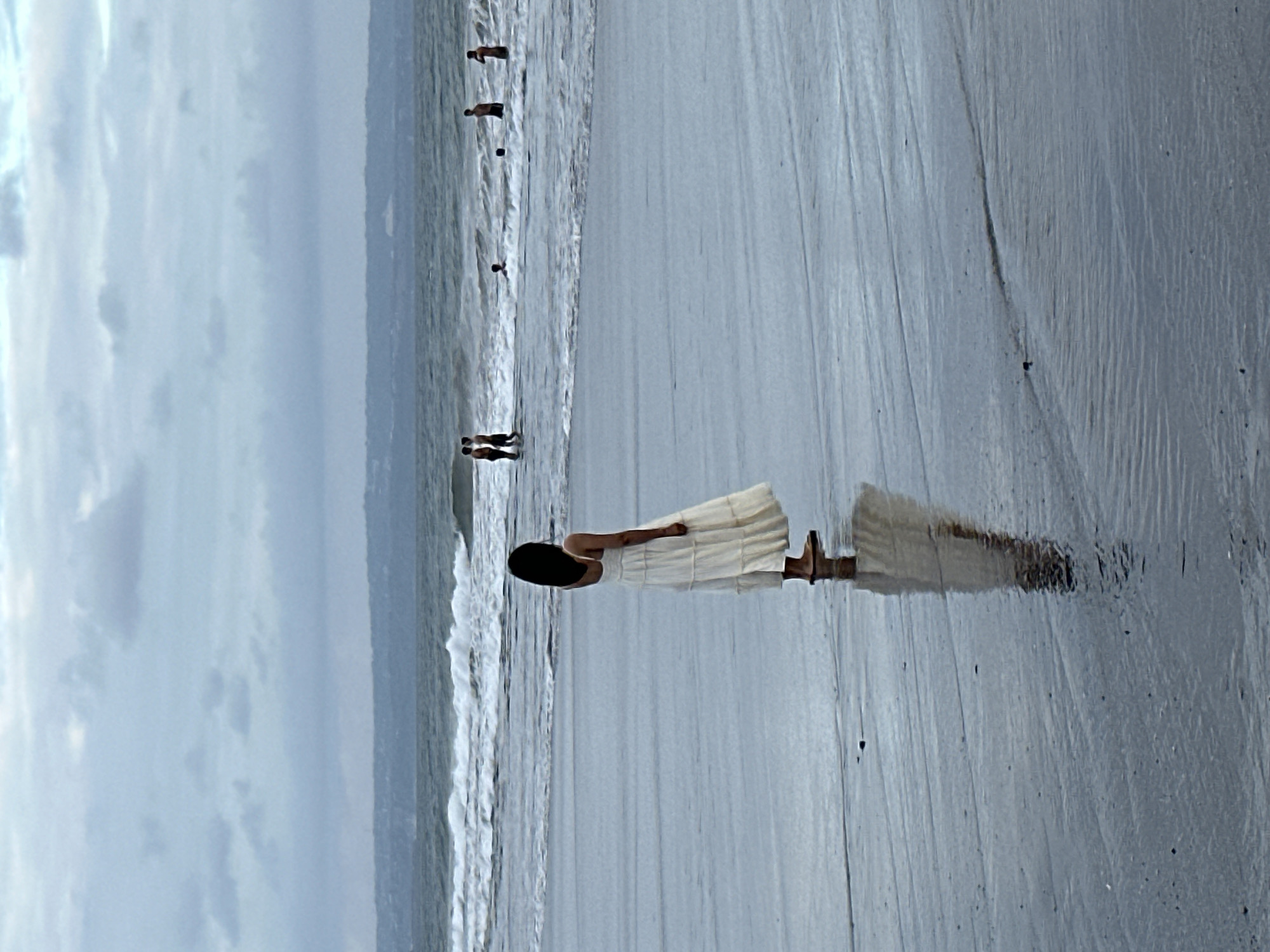 This image depicts a beach scene in North Kuta, Indonesia, under a cloudy sky. In the foreground, a woman wearing a long, white, tiered dress stands on the wet sand, facing away from the camera and towards the ocean. Her reflection is visible on the wet surface of the sand. In the middle ground, several people are in the water, some appearing to be swimming or wading. The ocean has moderate waves breaking on the shore. The background shows a hazy, distant coastline. The overall atmosphere is muted and overcast.