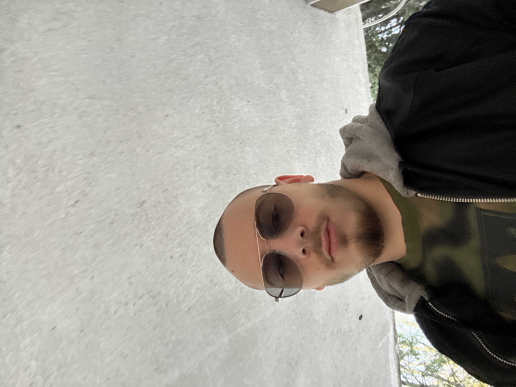 A man is seen in a selfie-style portrait, wearing sunglasses, a gray hooded sweatshirt, and a black jacket. He has a shaved head and a beard. The background appears to be a textured white wall. Beyond the jacket and wall, there is a suggestion of trees and sky. The exact location in Stara Zagora, Bulgaria is not discernable from this image.