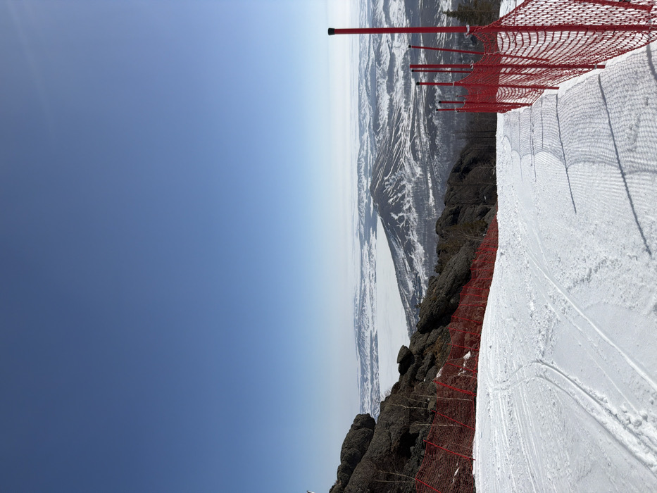 This image depicts a snow-covered ski slope descending from an elevated position in Zelenaya Polyana, Russia, under a clear, bright blue sky. The foreground shows groomed snow with distinct corduroy patterns and several ski tracks. To the left of the slope, dark, rugged rock formations are visible, interspersed with sparse, bare trees, and bordered by a red safety net that extends downwards. On the right side of the slope, a similar red safety net, supported by multiple red poles, marks the edge of the ski run. The mid-ground and background reveal a panoramic winter landscape featuring snow-covered mountains and hills, with a large, frozen body of water (likely a lake) visible as a flat, white expanse in the distance, partially shrouded by atmospheric haze. No individuals or active interactions are visible within the scene.