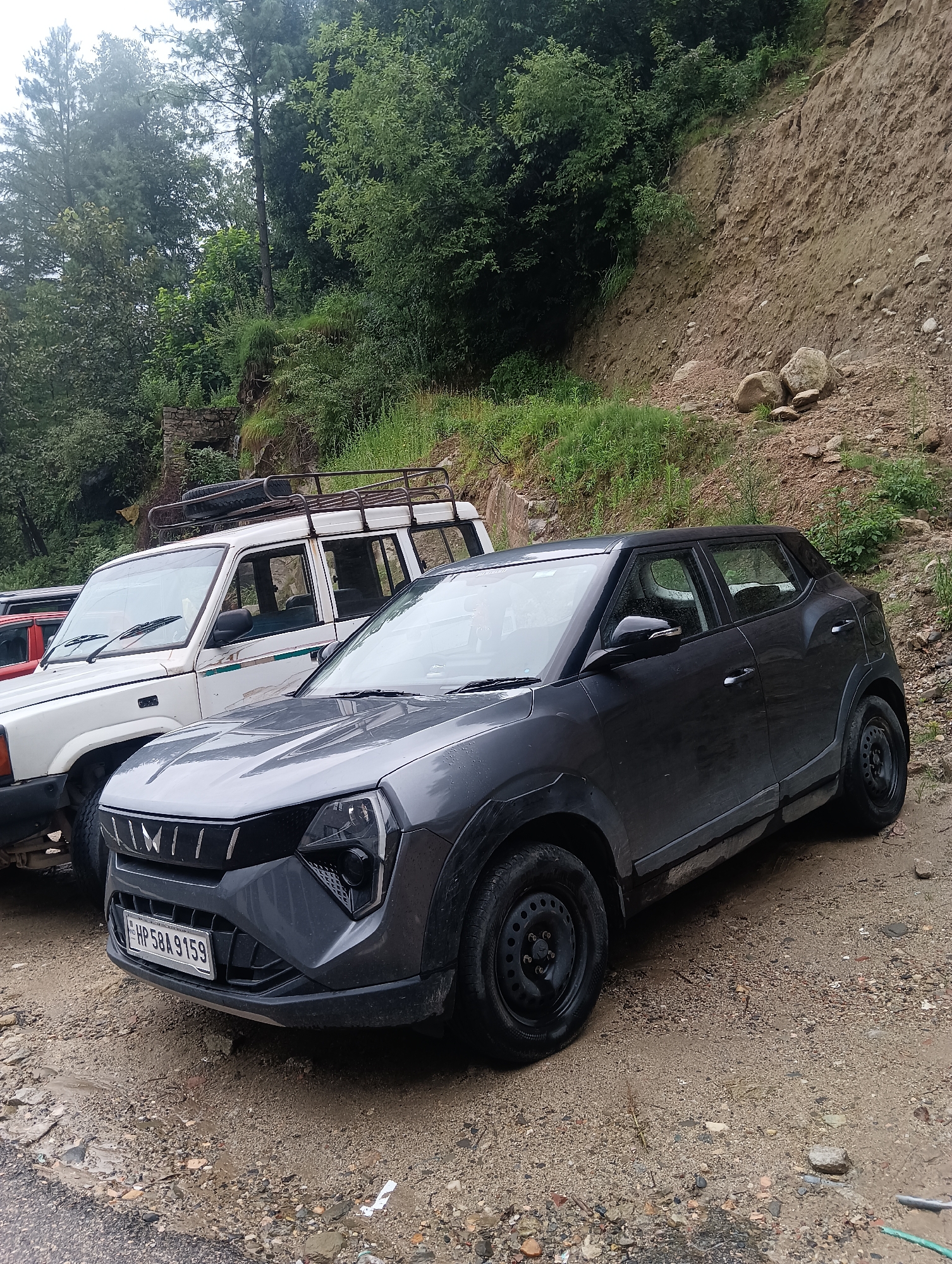 The image depicts a parked scene in what appears to be the city of Goshal, India, characterized by a dirt and gravel roadside adjacent to a steep, eroded hillside covered in vegetation and rocks. Two vehicles are visible: a dark gray SUV in the foreground and a white SUV with a roof rack behind it. The gray SUV, possibly a Mahindra XUV300, has its front license plate visible, reading "HP 58A 9159." The white SUV, possibly a Tata Sumo, has a spare tire mounted on its roof rack. The environment suggests a rural or mountainous area, with lush green trees and foliage forming a backdrop against the exposed soil of the hillside. The ground surface is uneven and wet, with puddles indicating recent rainfall. No people are present in the image.