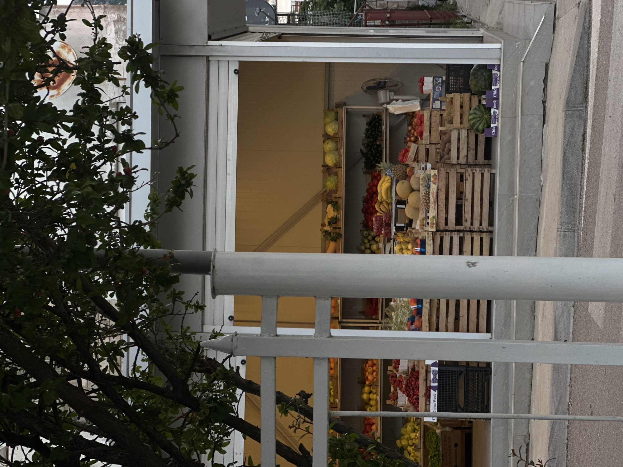 The image shows an outdoor fruit and vegetable stand in Šušanj, Montenegro. The stand is constructed with white siding and an open front displaying produce arranged in wooden crates. Fruits and vegetables, including melons, pineapples, bananas, tomatoes, lettuce, and other unidentified items are visible. A white railing partially obstructs the view of the stand, and a tree with green foliage is situated to the left. The street and sidewalk are visible on the right side of the frame.