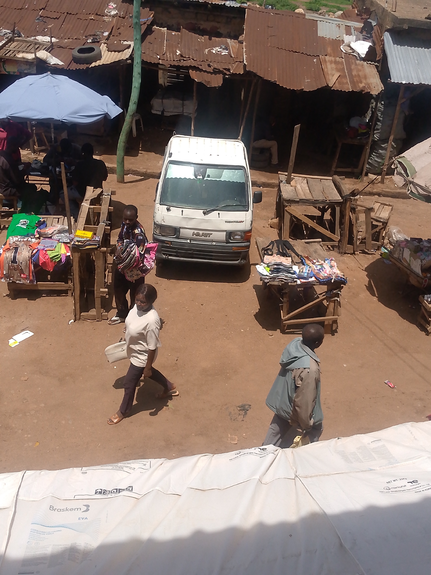 The image shows a busy street scene in Bukuru, Nigeria. In the foreground, part of a large white Braskem branded bag is visible. Further into the frame, there are several people walking and interacting with market stalls. A woman in a white top and dark pants walks in the foreground. Nearby, a man in a patterned shirt stands by a stall displaying colorful clothing items. A white van marked "HILJCT" is parked in the center of the scene. Other stalls display various goods. The buildings have corrugated metal roofs, suggesting a simple, perhaps informal, commercial area. An umbrella provides shade for a group of people sitting in one corner. The ground is mostly dirt.
