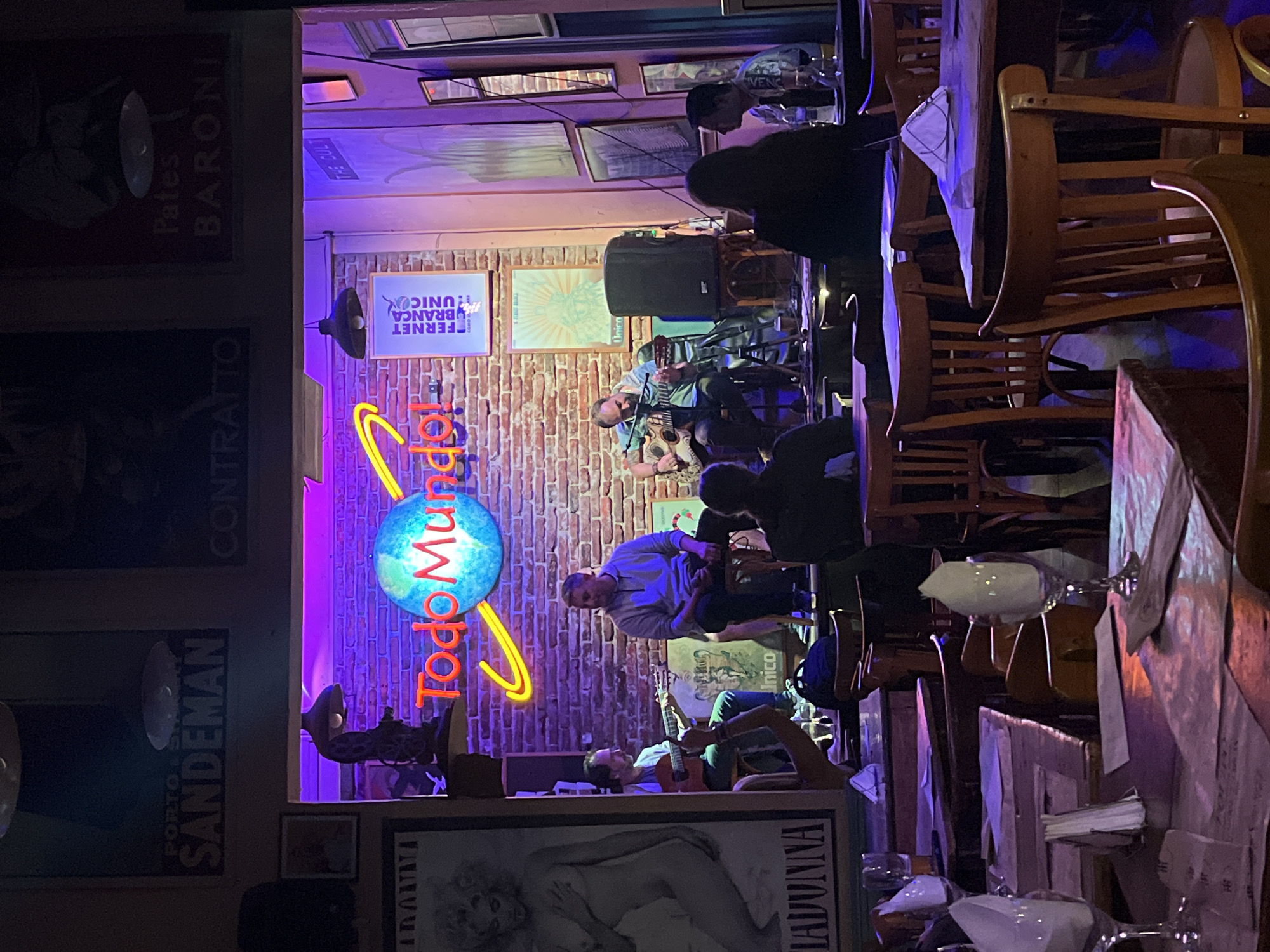 The photograph, taken at night and seemingly from the perspective of a diner at a table, depicts a bar/restaurant interior, likely in Buenos Aires, Argentina, given the Portuguese, English, and Spanish textual elements in the posters and neon sign "Todo Mundo."
A live band is performing on a small stage in the background, the band members are illuminated by purple lights. A brick wall serves as a backdrop, adorned with framed pictures and posters. A neon sign with the words "Todo Mundo" and illuminated rings around a globe is above the band. The foreground is dominated by tables and chairs, partially obscured by the angle and closeness of the shot. Glasses are set on the tables. Posters related to alcohol brands (Sandeman, Fernet Branca) are also visible.