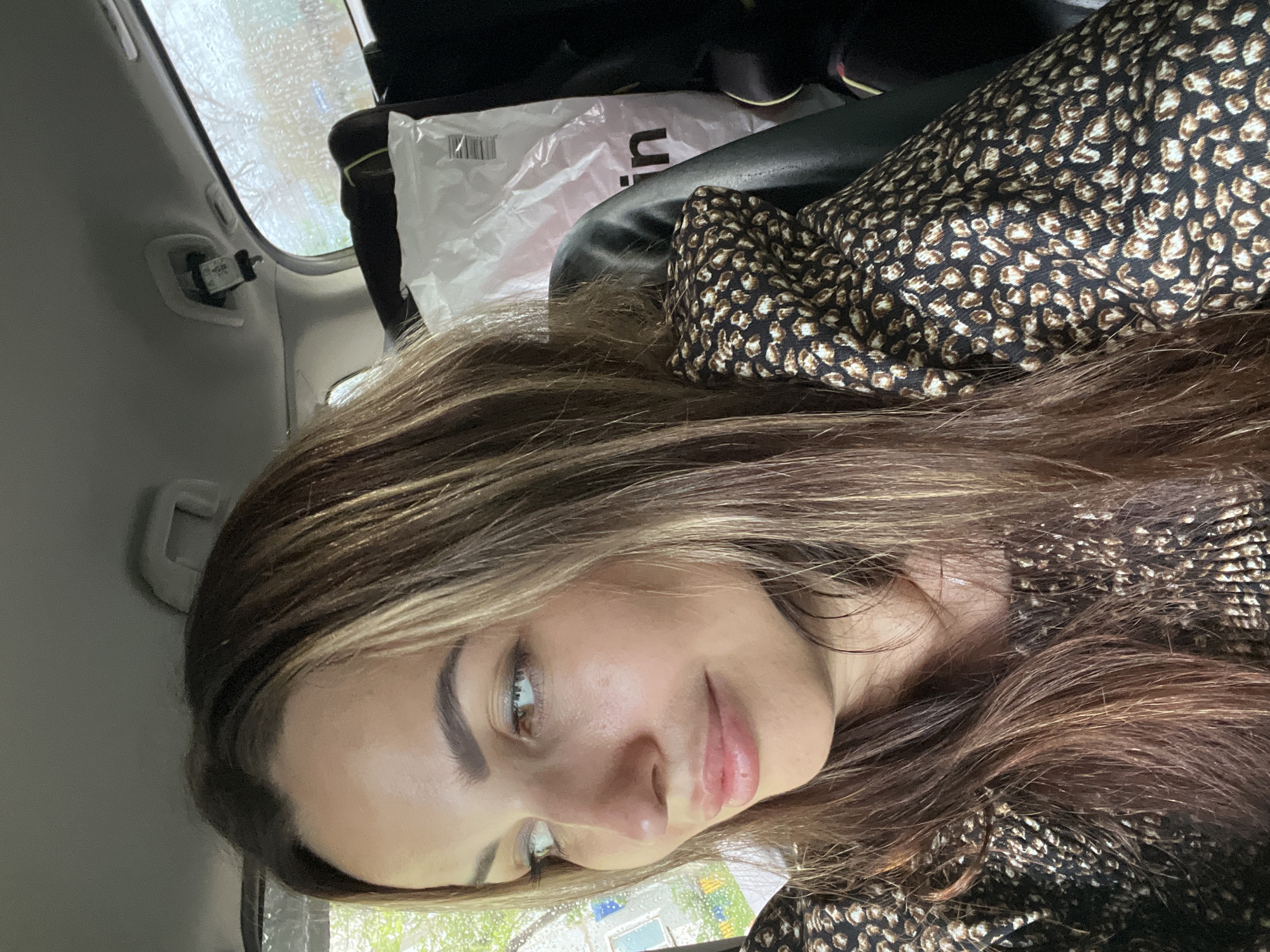 A woman with long, brown, highlighted hair is sitting in the backseat of a car. She is wearing a black and brown animal-print top. Her head is turned to the left, and she is looking off-camera. Raindrops are visible on the car window behind her, suggesting it is raining outside. A white plastic bag with lettering is behind her.
