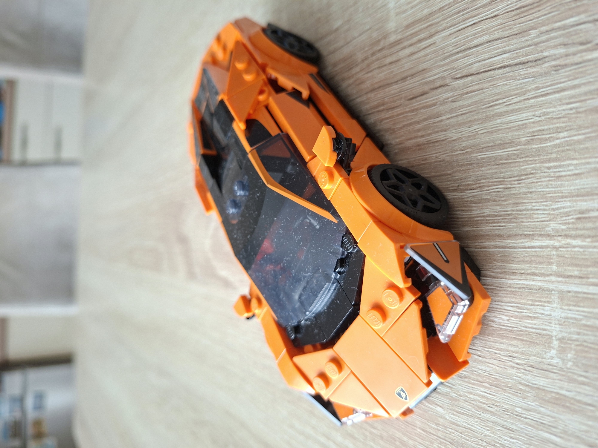An orange LEGO Technic Lamborghini Sián FKP 37 toy car is placed on a light brown wooden table. The car is facing to the right and slightly towards the viewer. Its headlights, windshield, and roof are black, and it has black wheels. The surface of the table has a visible wood grain pattern. The background is blurred, showing indistinct furniture that suggests an indoor setting. No people are present in the image, and no specific location in Baden-Baden, Germany is identifiable. The scene is static and well-lit.