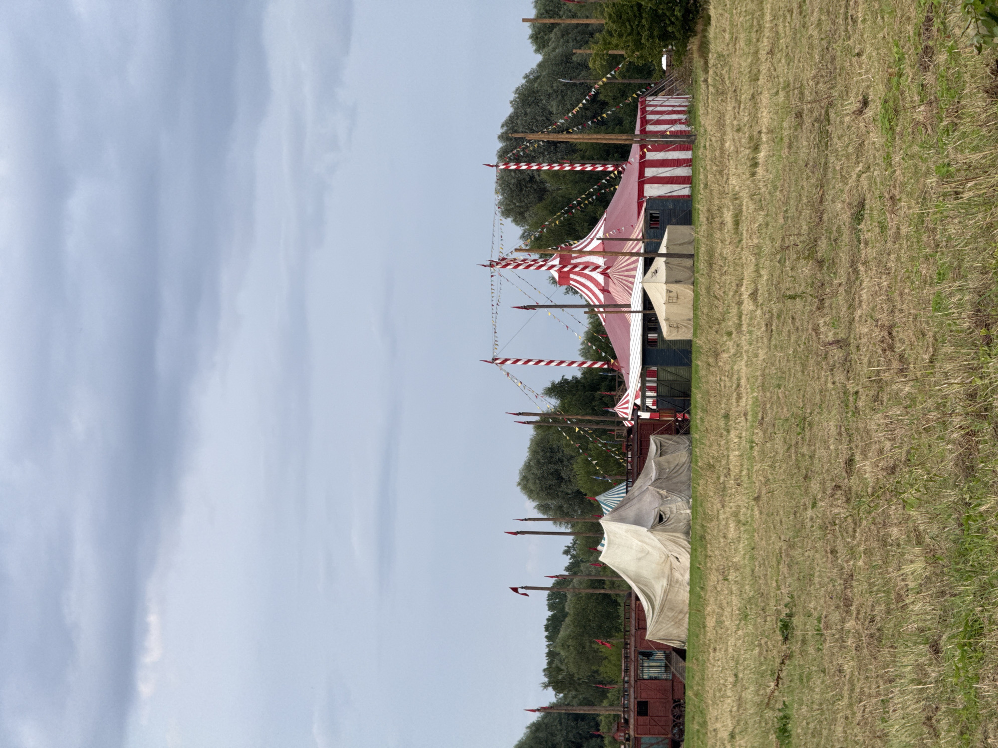 The image shows a rural outdoor scene with circus tents. In the foreground, there is a field of dry, brown grass. In the middle ground, there are several circus tents. The largest tent is red and white striped with a pointed roof. To the left of this tent, there is a large, off-white canvas tent, and to the left of that, a red train car. Behind the tents, there is a dense line of green trees. The sky above is overcast with gray clouds. The scene is not located in the city of Riga, Latvia; it is a generic rural setting.