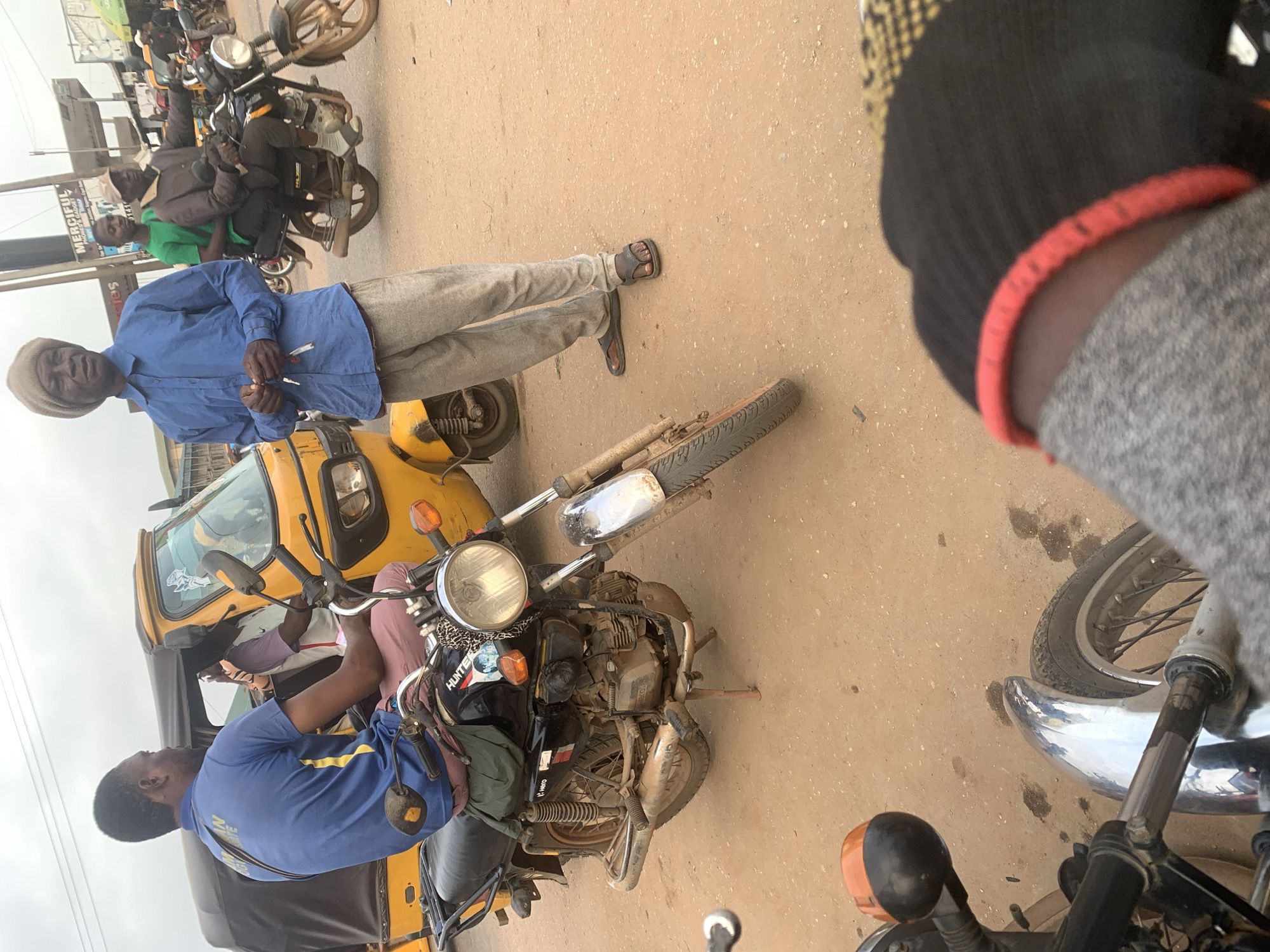 In Shagamu, Nigeria, a man in a blue long-sleeved shirt and beige trousers stands on a dusty road next to a yellow auto-rickshaw. He holds keys in his right hand. In the background, another man in a brown jacket and dark trousers rides a motorcycle, with a passenger in a green shirt behind him. Several other motorcycles and a motorcycle taxi are visible in the distance. The ground is dirt, and the sky is overcast.