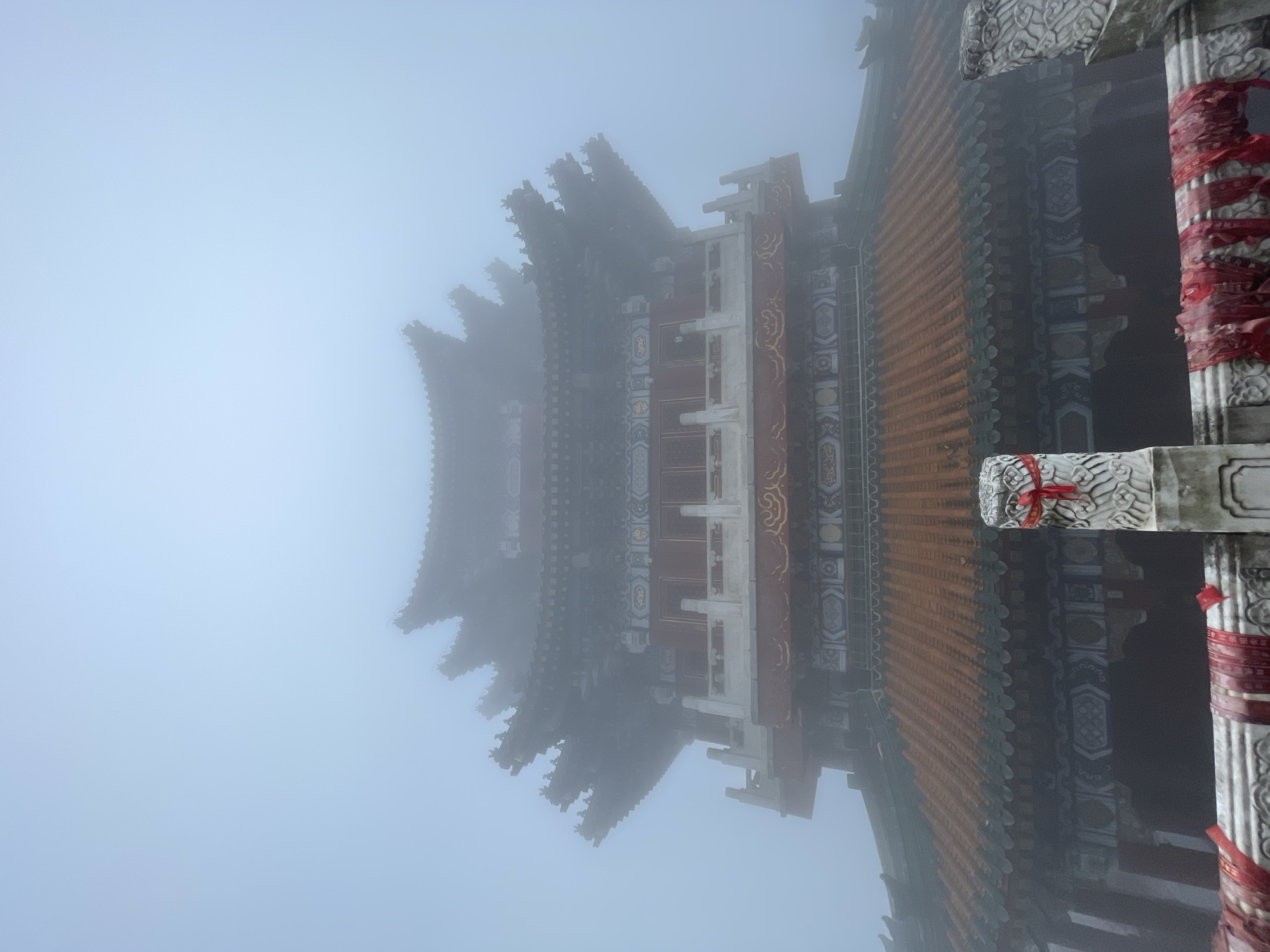 The image shows a portion of a traditional Chinese building shrouded in heavy fog. The building features a multi-tiered, ornate roof with upturned eaves. Details of the roof include orange-colored tiles and intricate carvings. The building's structure is visible, revealing red and white columns with decorative patterns. Red ribbons are tied around the columns. The fog obscures much of the building and surrounding environment. The photograph was taken in Yongding District, China.