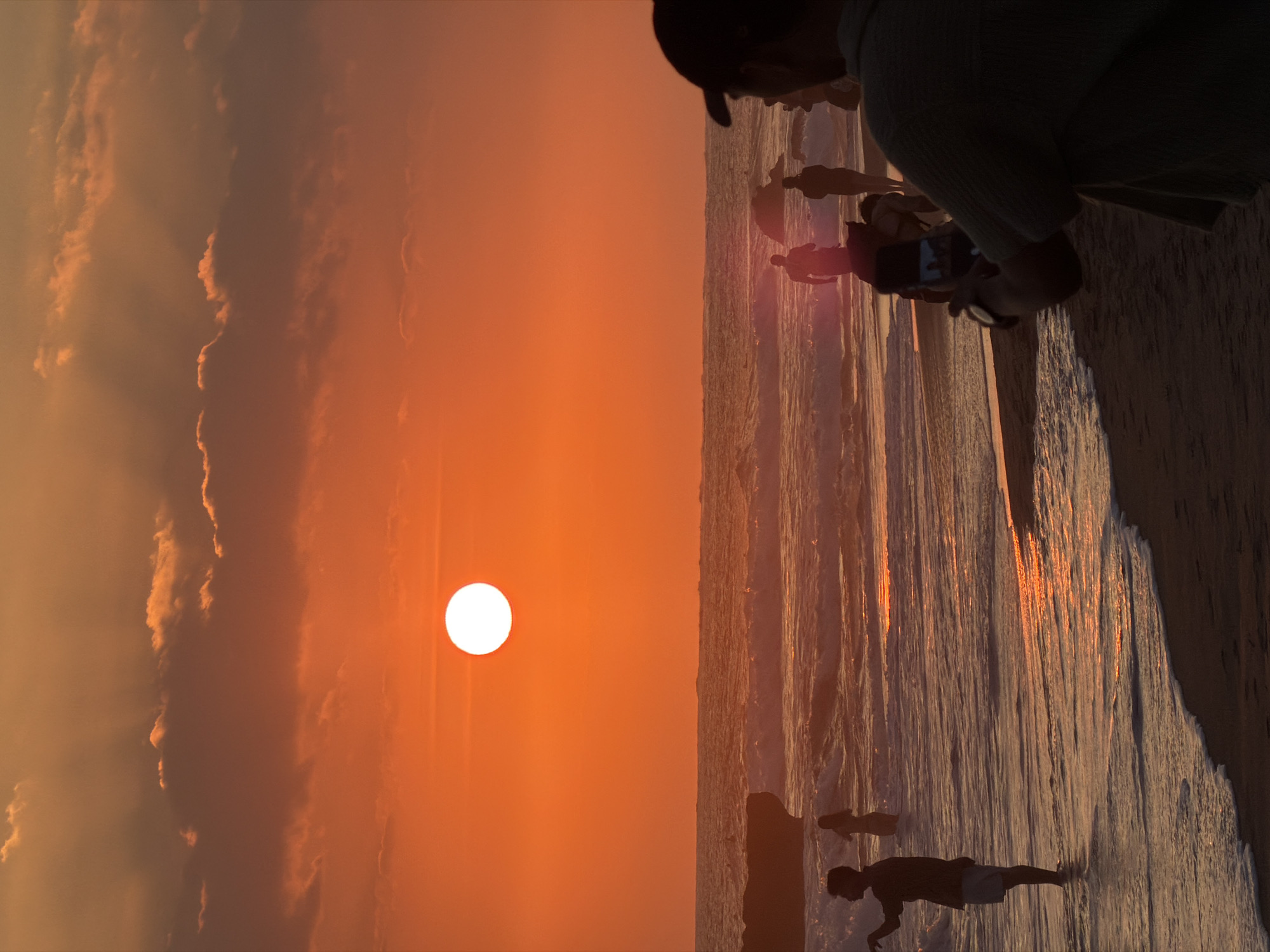 This is a vertical, eye-level, medium shot of a sunset at Echo Beach, North Kuta, Indonesia. The sun is a bright white orb in a vibrant orange sky, partially obscured by wispy, dark orange clouds. Rays of light emanate from the sun, illuminating the sky and reflecting on the water.
In the foreground, silhouetted against the sunset, a person wearing a dark baseball cap and a light-colored shirt is holding up a smartphone to take a picture or video of the scene. Their right arm is extended, holding the phone.
Further back, in the shallow water, several people are visible. On the left, a person in a dark shirt and light shorts stands facing the ocean. To their right, a person in a light-colored garment is visible in the water. In the mid-ground, a group of people are walking in the surf. Two figures, one darker and one lighter, walk towards the ocean. Behind them, more figures are present, some appearing to be holding umbrellas.
The ocean is calm, with gentle waves breaking on the shore. The water reflects the orange and red hues of the sunset. A dark, rocky outcrop is visible on the left side of the image, partially submerged in the water. The beach in the foreground is dark sand, with the edge of the water creating a foamy white line. The overall impression is a serene and picturesque sunset scene at a popular beach location.