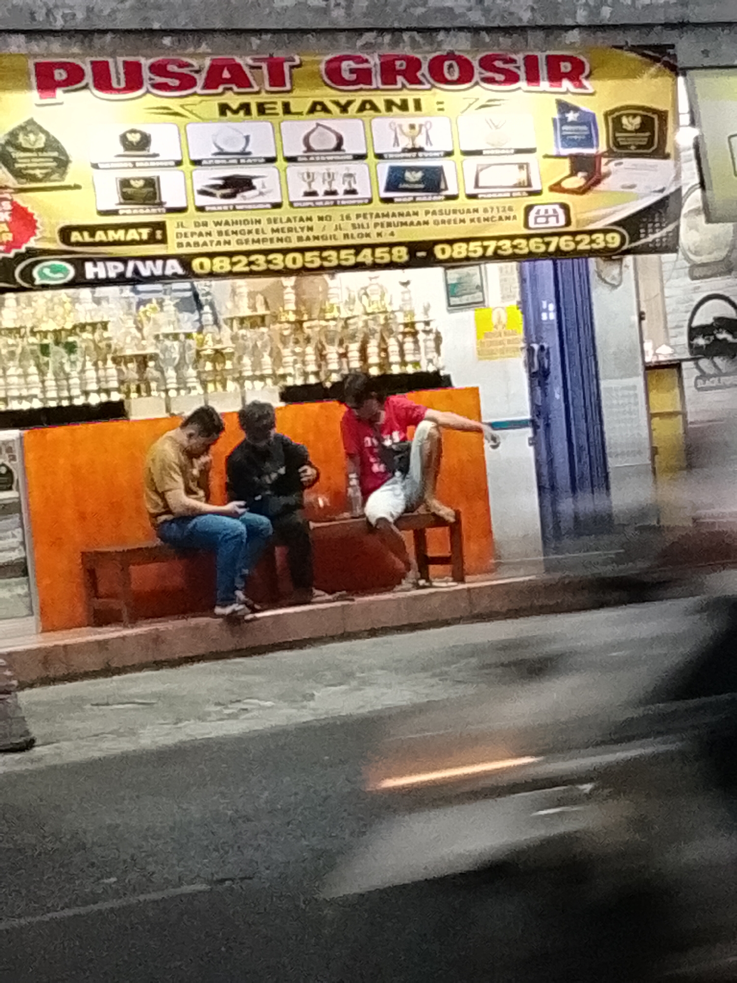 The image depicts an outdoor scene at night, likely on a street in Pasuruan, Indonesia, judging by the text on the banner. A large banner at the top advertises "PUSAT GROSIR MELAYANI," with smaller images of trophies, awards, and other items. Below the banner, three young men are seated on a wooden bench in front of an orange wall. The man on the left is wearing a yellow t-shirt and blue jeans, looking down at his phone. The man in the middle is wearing a black shirt and dark pants, also looking at his phone. The man on the right is wearing a red t-shirt and white shorts, with his legs stretched out. The background shows rows of trophies, suggesting the establishment is a trophy shop or a place that sells awards. The street in the foreground is wet, indicating recent rain.