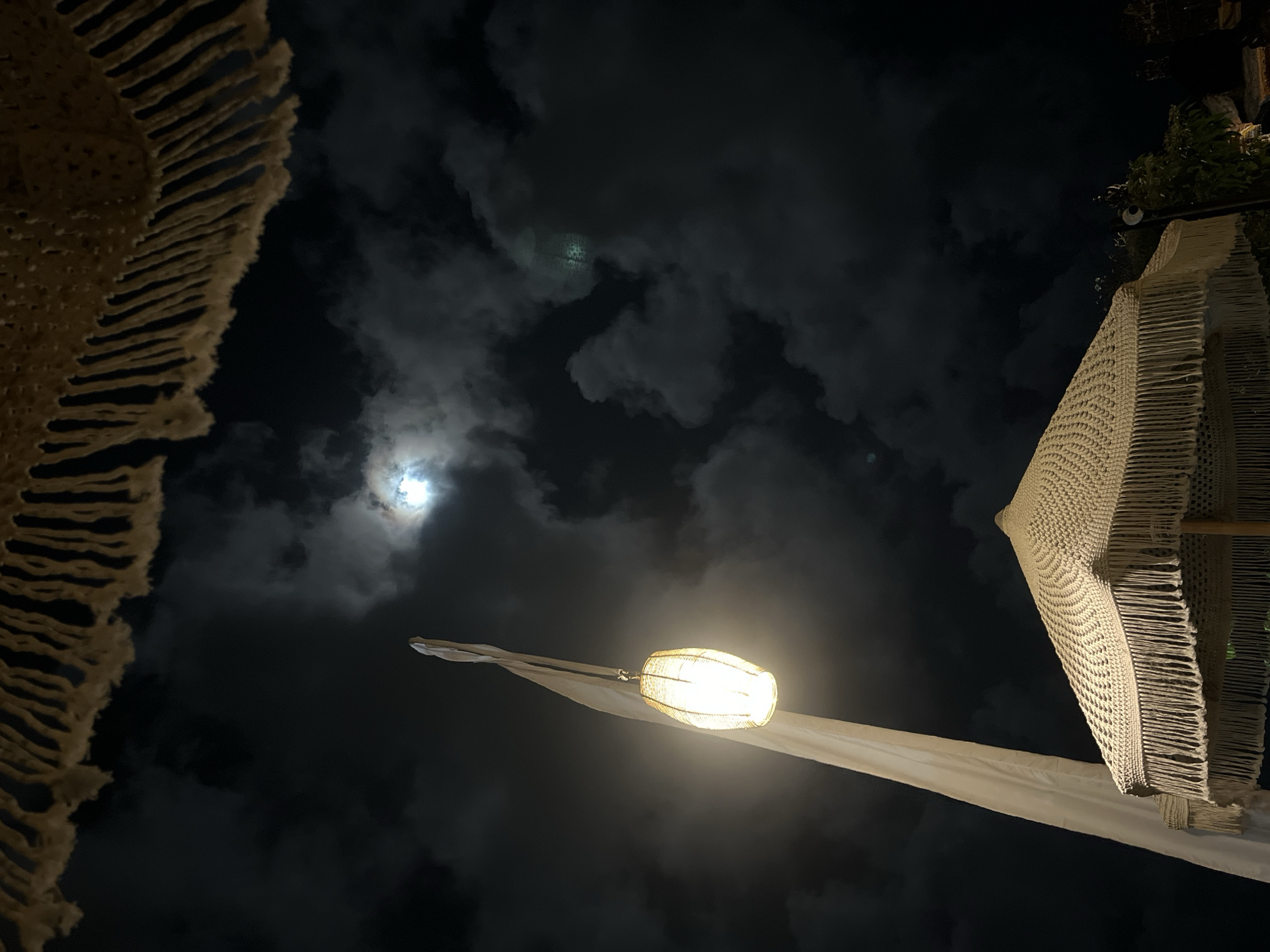 The image shows a nighttime scene in Kuta Selatan, Indonesia. The sky is dark, with a few clouds and a bright moon visible through them. In the foreground, there is a decorative light fixture suspended from a white fabric banner. Two white fringed sunshade umbrellas are partially visible on the left and right.