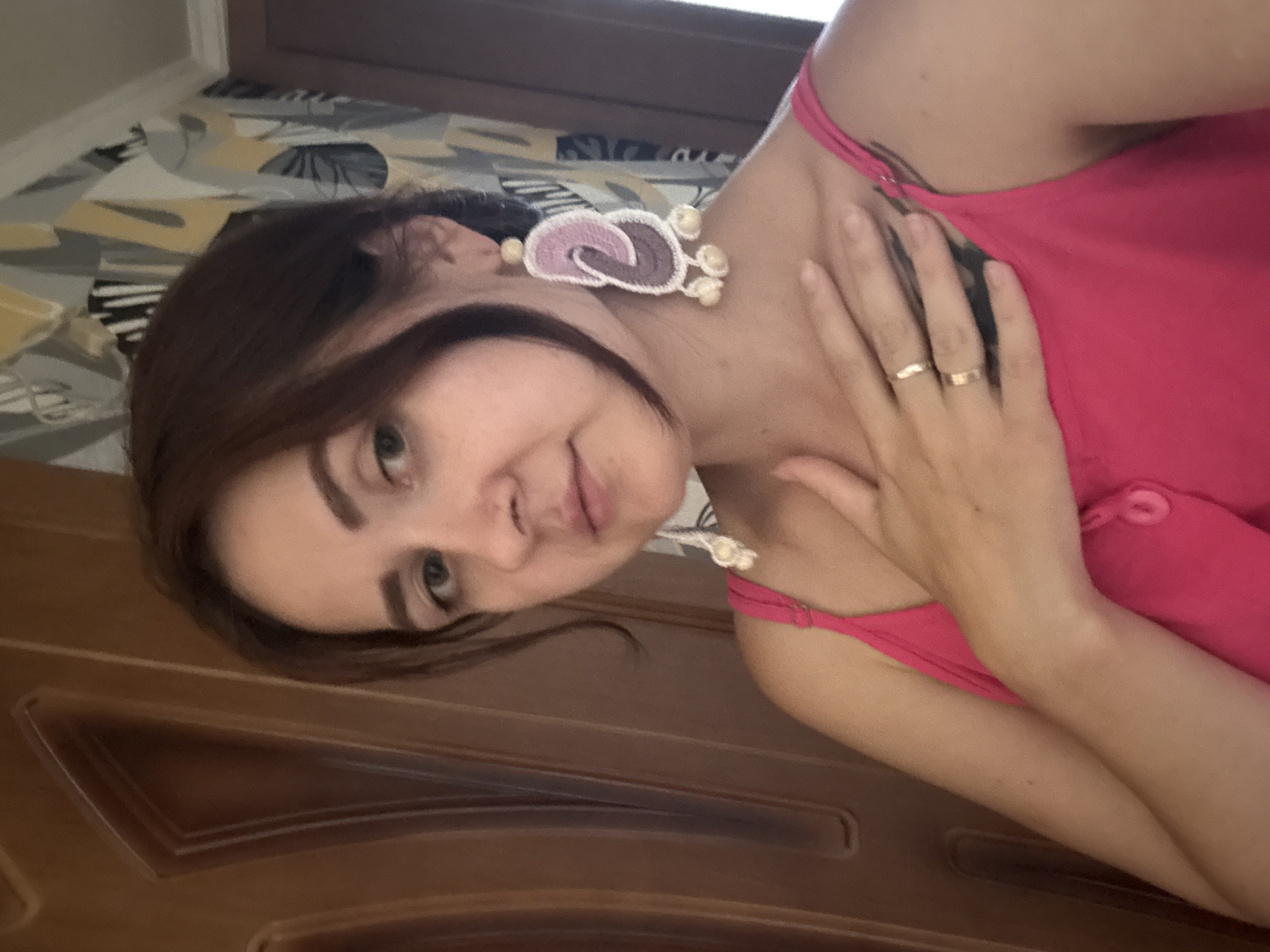 A young woman with brown hair and fair skin is looking into the camera, holding her hand to her chest. She is wearing a bright pink tank top and large, dangling earrings. The background appears to be an indoor setting with a wooden door on the left and patterned wallpaper on the right. The lighting suggests it is daytime. The image appears to be a selfie taken by the woman. There is no indication of the location being the city of Naberezhnye Chelny, Russia.