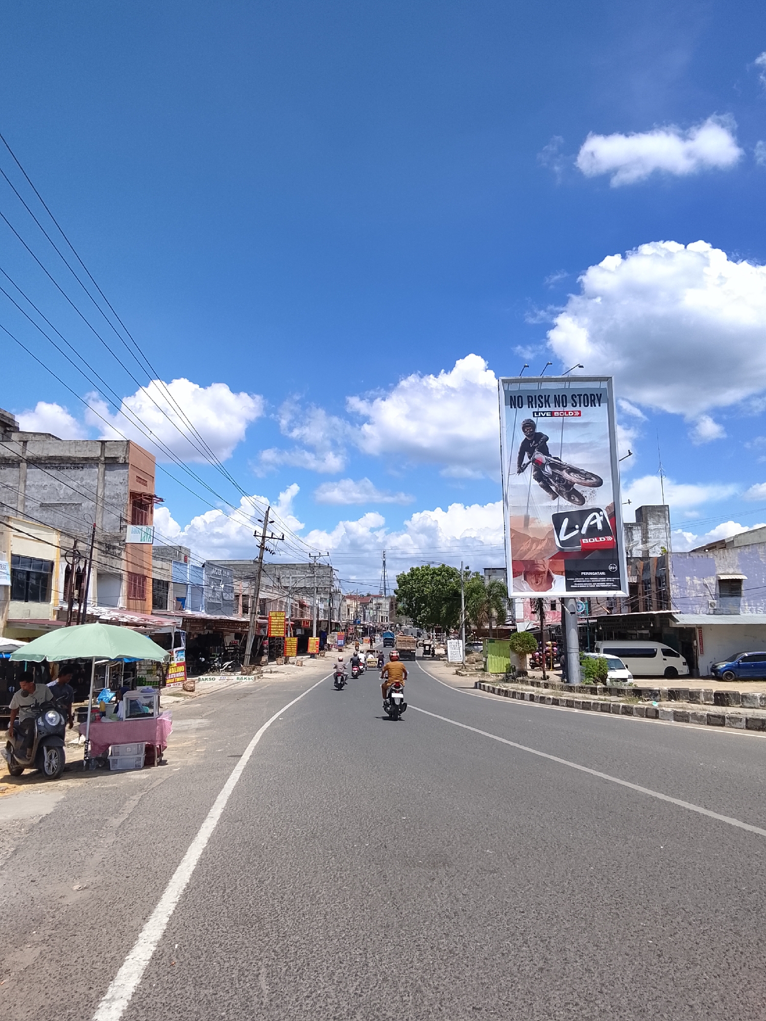 The image depicts a street scene in Lhokseumawe, Indonesia, under a bright, partially cloudy blue sky. The street is paved with asphalt and has a white dividing line. On the left side of the road, there are several buildings lining the street, some appearing older and weathered, with awnings and signs. A green umbrella shades a small food cart with a checkered tablecloth and a cooler. Several motorcycles are parked or in motion.
On the right side of the road, there is a large billboard advertising "LA BOLD" with an image of a person on a motorcycle. Further down the road, more buildings and businesses are visible. Several people are riding motorcycles on the street. A white van and a blue car are parked on the right side of the road. Power lines crisscross the sky above the street. The overall impression is a typical urban street scene in a tropical climate.