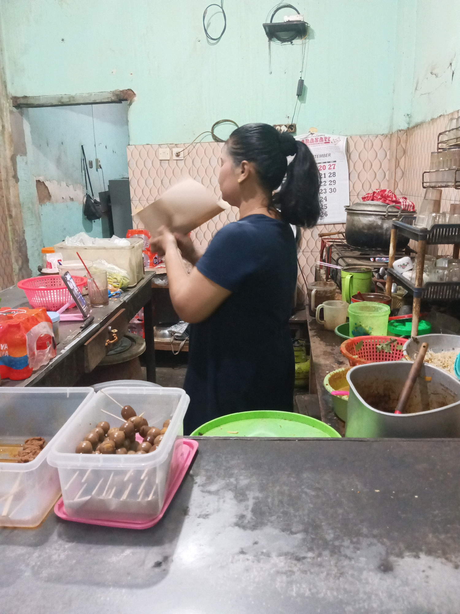 The image depicts a woman in a dark blue shirt holding a brown paper bag in what appears to be a food stall or kitchen in Kalangbret, Indonesia. The environment is cluttered and appears functional, with various containers of food, cooking utensils, and appliances visible. In the foreground, there are several plastic containers holding food items, including what looks like quail eggs on skewers in one. A metal counter runs across the bottom of the frame. The background shows a light green wall with some peeling paint and a calendar on the wall. A shelf unit with dishes and glasses is also visible to the right. The lighting is somewhat dim, suggesting an indoor setting.