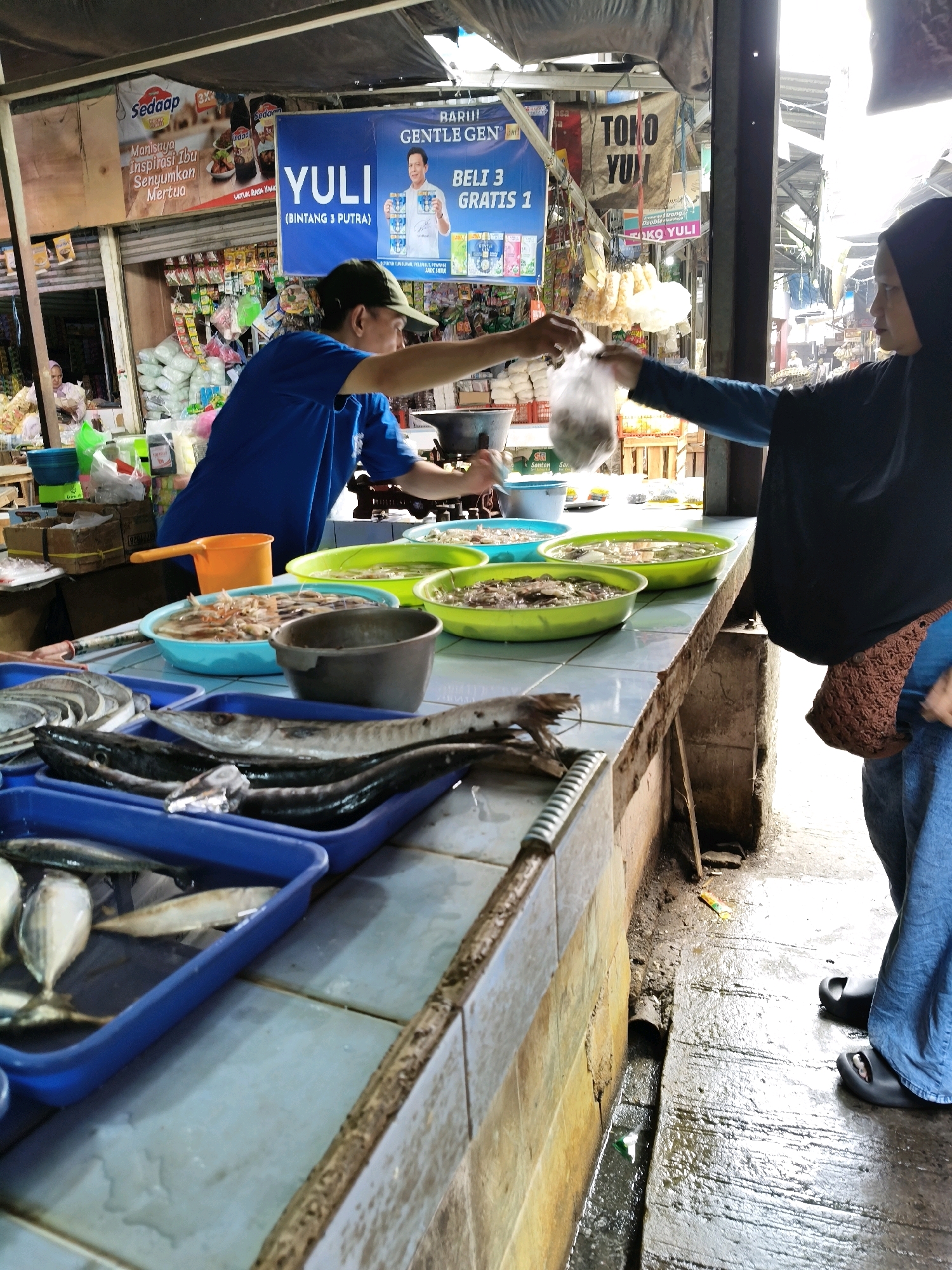 At a market stall in Setiamekar, Indonesia, a vendor in a blue shirt and green cap is handing a plastic bag filled with small items to a customer wearing a black hijab and blue pants. The customer's right hand is visible, holding the bag. The market stall has a counter displaying various types of fish in blue trays, including what appear to be mackerel and smaller fish. Behind the counter, several large, green, shallow bowls are filled with water and what looks like seafood, possibly shrimp. To the left of the customer, a brown woven bag hangs from their side. In the background, the market is partially covered by a black tarp, and signs advertising "SEDAP" and "YULI" are visible. The ground around the stall is wet, suggesting recent rain or cleaning, with a drainage ditch running along the base of the stall. The overall scene depicts a transaction at a fresh food market.