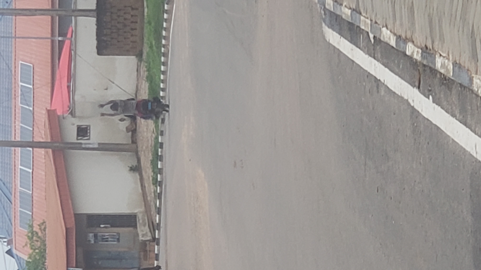 The image, taken in Narayi, Nigeria, depicts a street scene with a road running horizontally and buildings on the left side. The lighting suggests it is daytime.
On the right, a gray asphalt road occupies a large portion of the frame. A solid white line runs along its left edge, bordering a curb. This curb is distinctive with alternating black and white stripes.
To the left of the curb is an unpaved shoulder or sidewalk area. Here, two or three indistinct human figures are visible. They appear to be walking or standing. One figure, centrally located, seems to be wearing light-colored clothing on top and darker clothing below, possibly carrying an item or wearing a backpack. The features of the individuals are not clear due to distance and image resolution.
Further to the left, a series of buildings and structures are present. A prominent building features a reddish-brown sloped roof. Numerous solar panels are installed across this roof. Below the roof, a section of a light-colored wall is visible, with a small dark window cut into it. A bright red cloth or tarp is draped over part of the building or an adjacent structure near the roofline. Behind this building, there's another section of wall, partially constructed with reddish-brown bricks. Two dark vertical poles, possibly utility poles, stand in the foreground on the left side. Some sparse green grass and a small plant are also visible near the buildings and curb.
The image is somewhat blurry and grainy, particularly the distant figures and details. No legible text is visible in the frame.