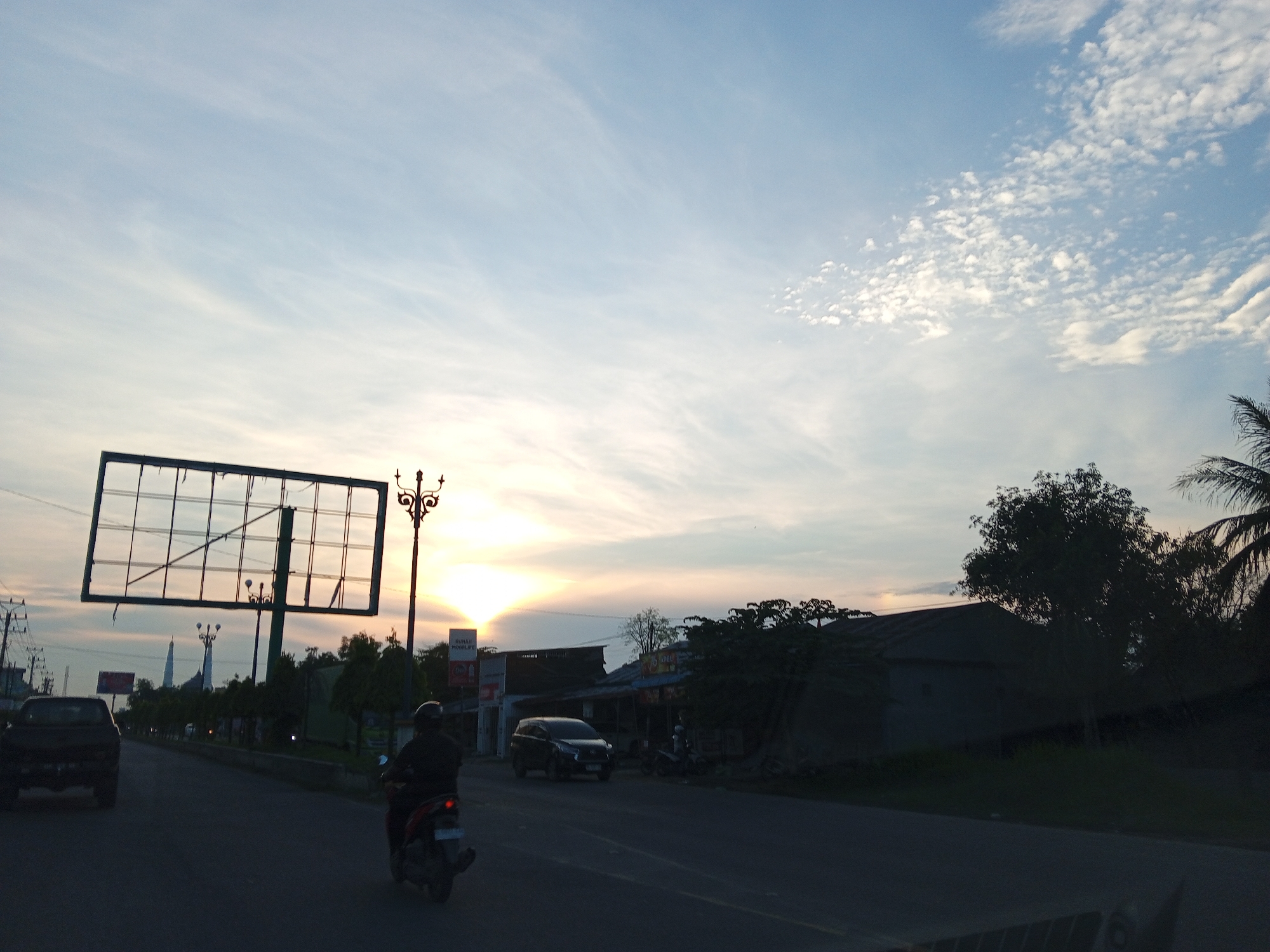 This is an outdoor, eye-level, medium shot taken at dusk in Pantegajah, Indonesia. The sky is filled with wispy clouds, illuminated by the setting sun which casts a warm glow on the horizon. In the foreground, a dark-colored car drives down a paved road. To the right of the car, a person on a red and black motorcycle is also traveling down the road. In the background, there are several buildings with dark roofs, trees, and utility poles. A large, empty billboard stands on the left side of the road, and a decorative street lamp is visible to its right. The overall scene depicts a quiet street in a semi-urban area during twilight.