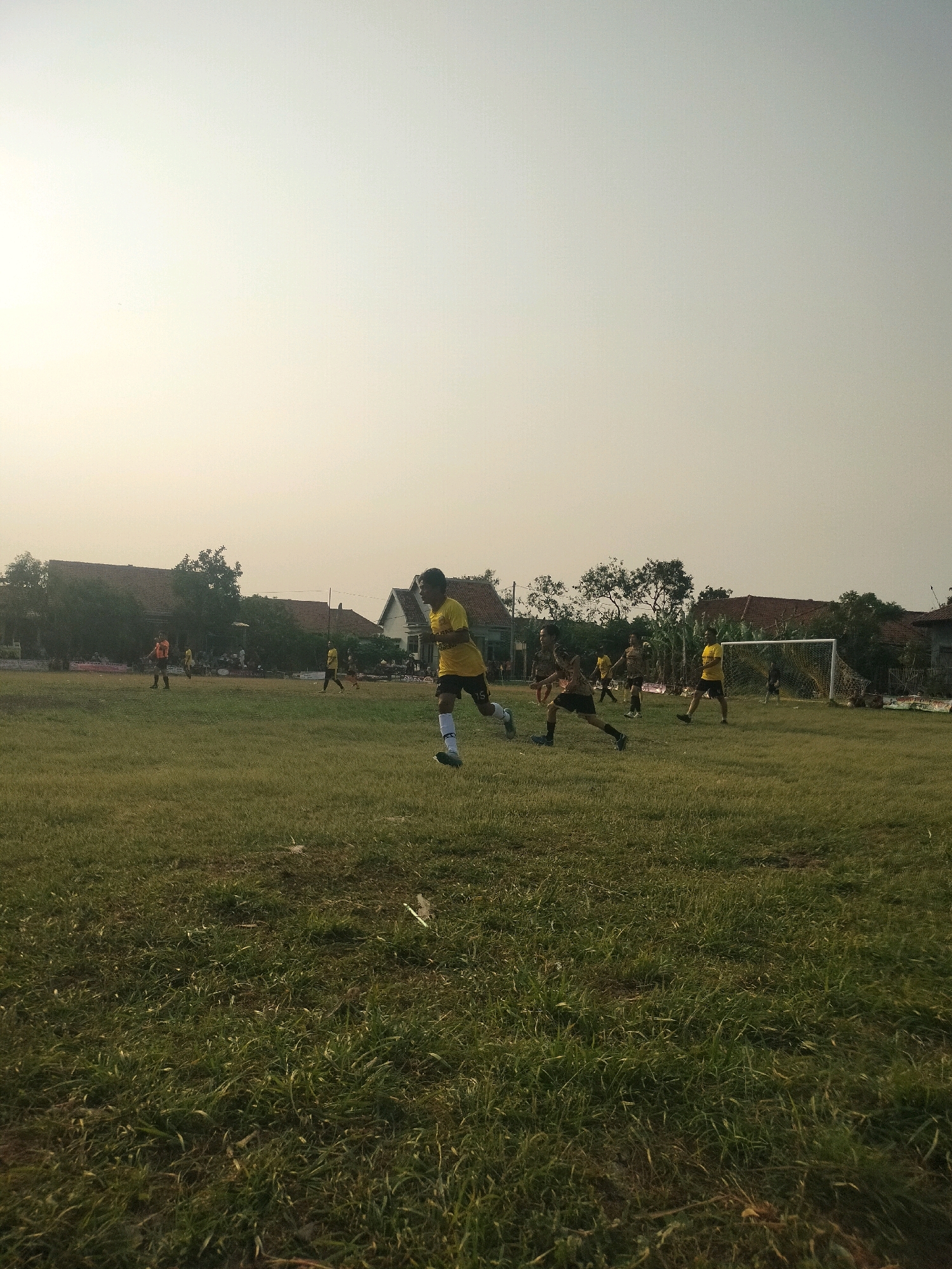 The image depicts a youth soccer game taking place on a grassy field in Kalisari, Indonesia. The sky is overcast, and the lighting suggests it is either early morning or late afternoon. Several young boys, mostly wearing yellow jerseys, are actively engaged in the game. In the foreground, a player in a yellow jersey is running towards the right of the frame, his body angled as if in motion. Other players are visible in the mid-ground, some running with the ball, while others are positioned defensively. A soccer goal with a white net is situated on the right side of the field. In the background, houses and trees are visible, indicating a residential area. The overall scene captures a moment of athletic activity and community engagement within the village.