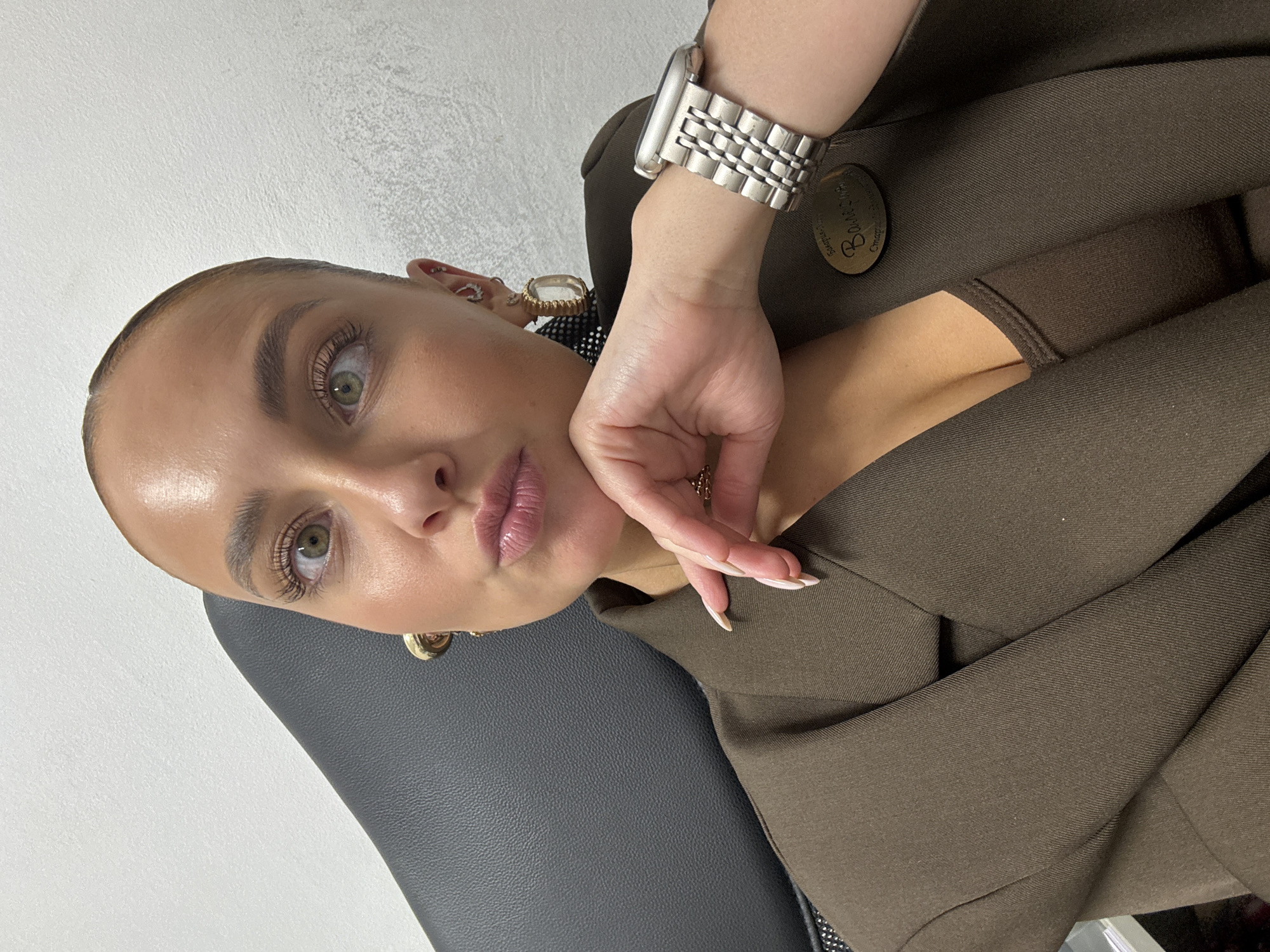 A female subject is captured in an indoor selfie, positioned in what appears to be a dark grey upholstered chair. Her hair is pulled back, revealing her forehead and prominent green eyes. Her lips are puckered in a pouting expression, and her right hand, featuring long, French-tipped fingernails and a gold-toned ring, rests under her chin. She is wearing large, gold-toned, abstract-shaped earrings in both ears and multiple silver-toned hoops and studs in her left ear. A silver-toned Apple Watch with a metallic link band is on her left wrist. Her attire consists of a dark brown blazer over a similarly colored V-neck top. A name badge pinned to her right lapel displays "Валерия" (Valeria) and "Старший Специалист" (Senior Specialist) in Cyrillic. The background is a plain, textured, light grey wall. No specific architectural or geographical features are present to identify a precise location within the city of Chaikovsky, Russia.