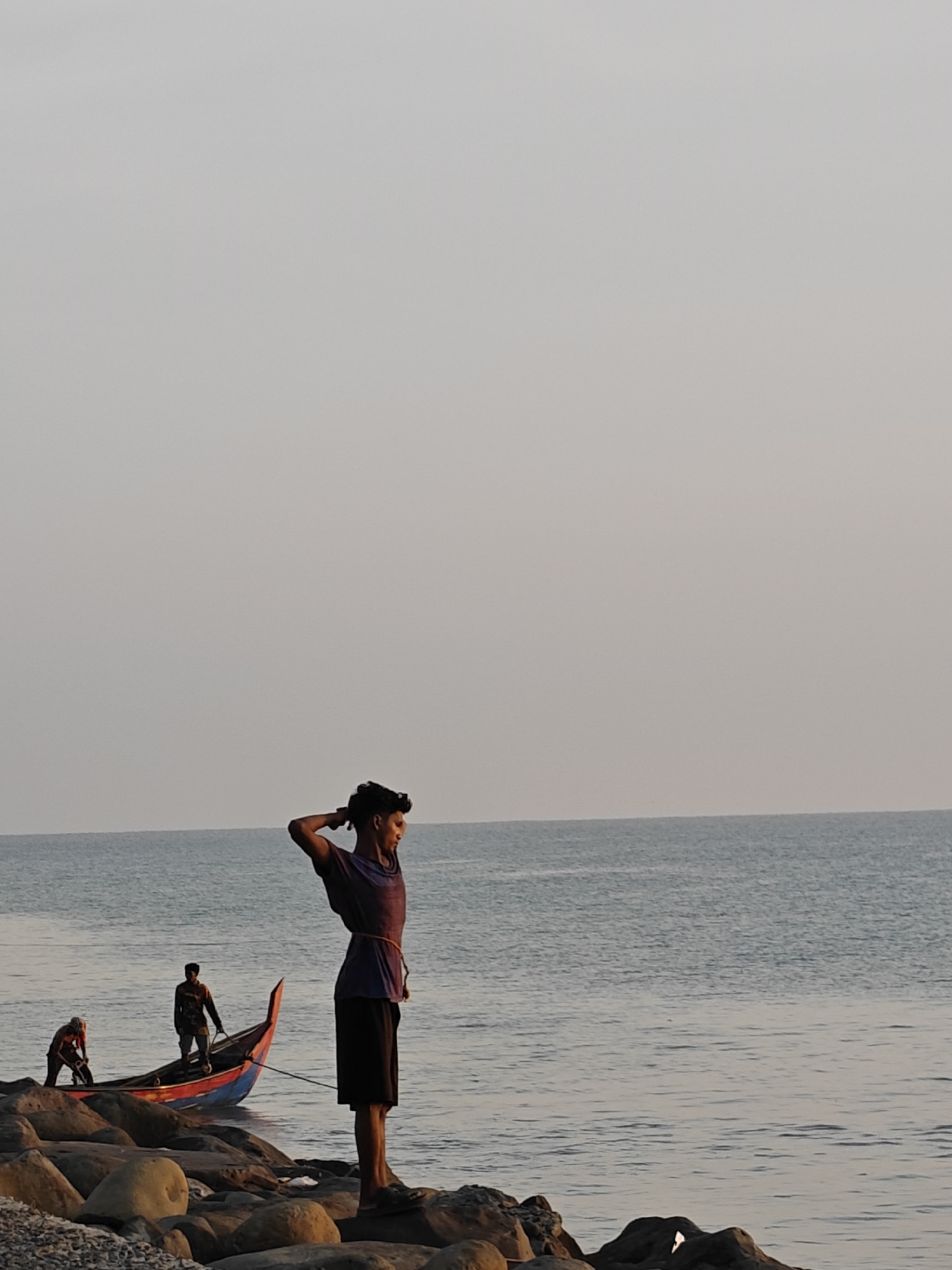This image depicts a serene coastal scene in Lhokseumawe, Indonesia, likely captured during sunrise or sunset, given the soft, diffused lighting. The expansive sky is a pale, uniform gray, meeting a calm, light blue ocean at the horizon.
In the foreground, a young man stands on large, dark rocks that form part of the shoreline. He is positioned towards the right side of the frame, looking out over the water with his hands clasped behind his head, suggesting a moment of reflection or relaxation. He has dark, curly hair and wears a dark t-shirt and dark shorts.
Further back, closer to the water, a brightly colored traditional wooden fishing boat with red and blue paint is visible. Two other men are engaged with the boat: one stands inside it, while another, wearing a reddish-orange top, is near the bow, appearing to work with the vessel, possibly preparing it for launch or securing it ashore. The rocks continue along the water's edge, creating a rugged coastline. The scene conveys a sense of peaceful daily life and connection to the sea within a coastal community. There is no visible text in the image.