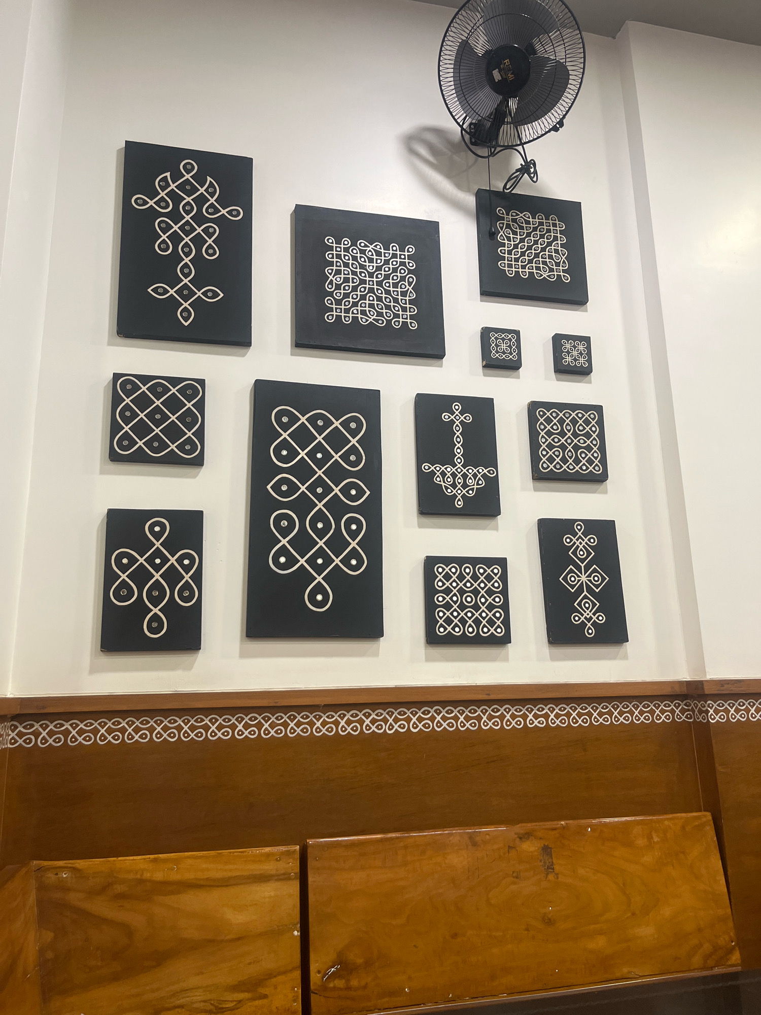 The image shows a wall in a building in Navrangpura, India, decorated with several framed Kolam designs. The Kolams are geometric patterns created with white lines and small circles on black backgrounds. There are various sizes of framed Kolams arranged in a grid-like fashion on the wall. A black oscillating fan is mounted on the wall above the Kolams. The lower part of the image shows wooden benches and a decorative border with a repeating pattern along the bottom of the wall.