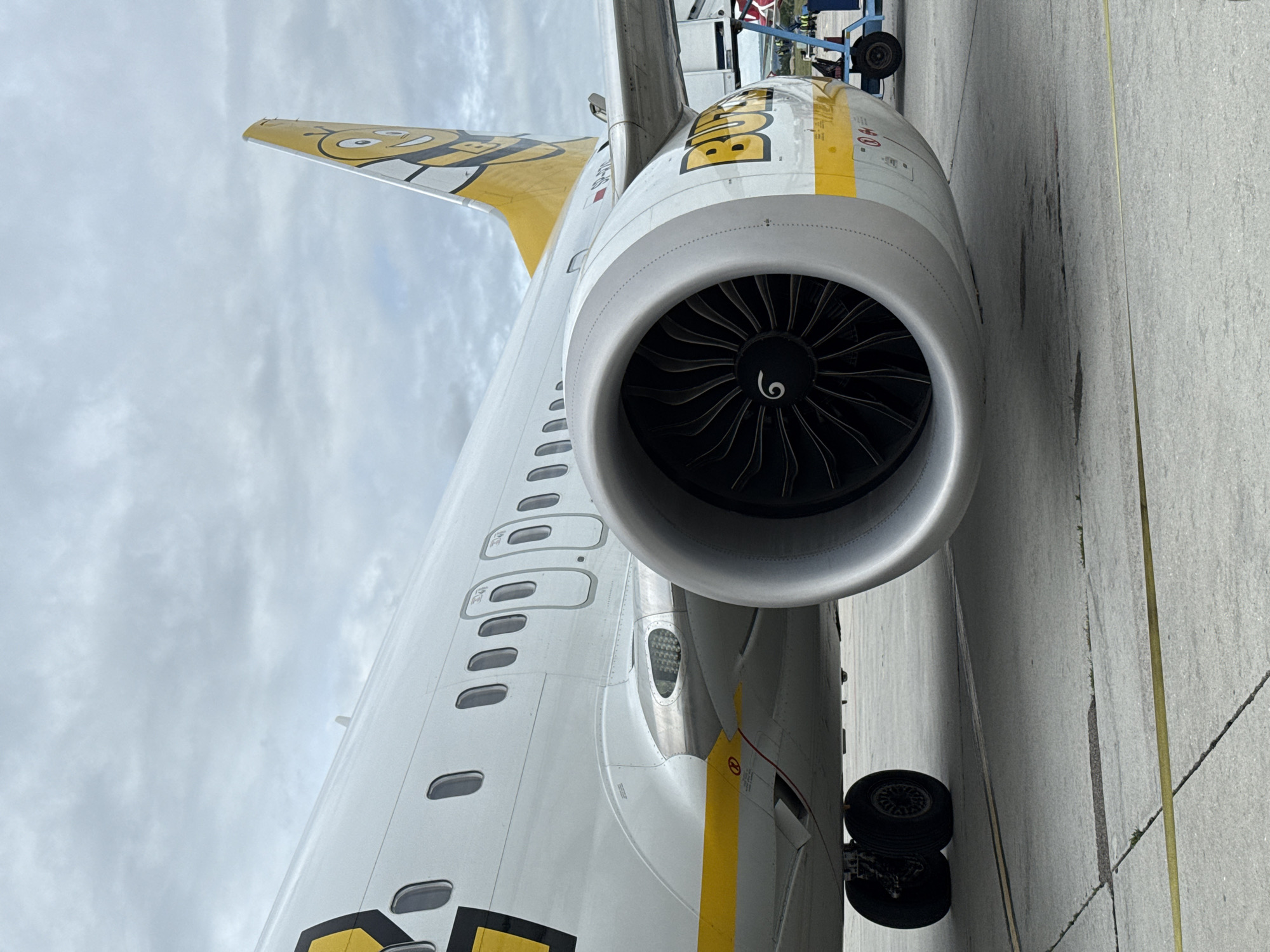 The image shows a close-up of an airplane engine and the side of the fuselage, resting on a tarmac. The aircraft is white with yellow accents and features a cartoon bee logo on the tail. The engine is large and silver with black fan blades visible inside. There are several windows visible along the side of the plane. The tarmac is concrete with yellow lines. In the background, there is a cloudy sky and some airport equipment, including a blue baggage cart. The plane appears to be parked at an airport. It is not possible to determine the specific location in Aksakovo, Bulgaria from this image.