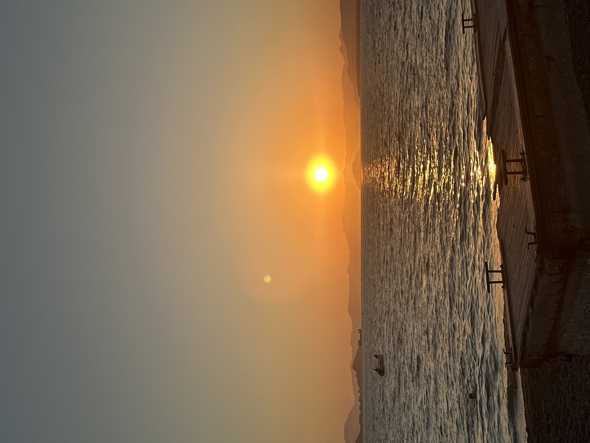 A vertical, full-shot photograph captures a serene sunset over the sea, likely in Fethiye, Turkey, given the distinctive mountainous coastline. The sky is a gradient of soft peach and orange hues, transitioning into a dusky blue at the very top. The sun, a bright, intense yellow orb, is positioned slightly left of the center, casting a shimmering, golden reflection across the gently rippling water.
In the mid-ground, a small boat with at least two people is visible, navigating the calm sea. To the far left, a person's head and shoulders are just visible above the water, suggesting they might be swimming.
The foreground is occupied by a wooden pier or dock, constructed from weathered planks. Two small, dark, bench-like structures are positioned on the pier. The pier appears to be at the edge of a pebble beach, with dark, coarse stones forming the shoreline. In the background, a chain of layered, dark mountains lines the horizon, silhouetted against the vibrant sunset. The overall atmosphere is peaceful and tranquil.