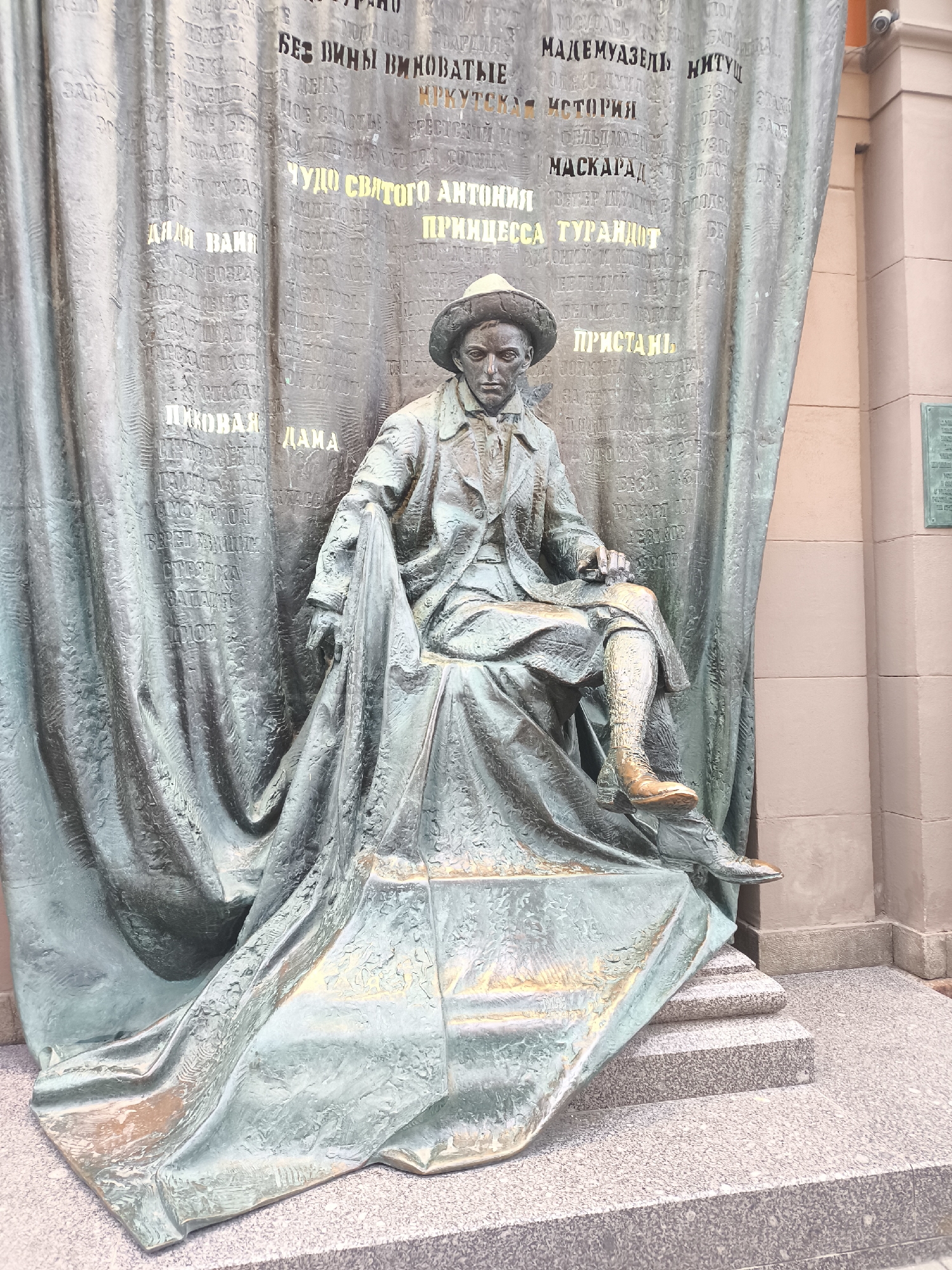 A bronze monument, likely depicting a playwright or theater director, is captured in an outdoor setting in Moscow, Russia. The central subject is a male figure seated in a relaxed, contemplative pose, wearing a hat, a coat, and trousers, with one leg crossed. His right shoe exhibits a distinct polished sheen, indicating frequent touching by visitors.
Behind the seated figure is a large bronze backdrop sculpted to resemble a draped theater curtain. This curtain is intricately textured and features numerous Russian words and phrases inscribed across its surface, many highlighted in a brighter, possibly gold, tone. These inscriptions are titles of prominent plays and literary works, including "Без вины виноватые" (Without Guilt, Guilty), "Мадемуазель Нитуш" (Mademoiselle Nitouche), "Иркутская история" (An Irkutsk Story), "Маскарад" (Masquerade), "Чудо святого Антония" (The Miracle of Saint Anthony), "Принцесса Турандот" (Princess Turandot), "Пристань" (The Pier), "Пиковая дама" (The Queen of Spades), and "Дядя Ваня" (Uncle Vanya).
The monument rests on granite steps, with a building wall visible to the right. The overall bronze material displays varying patinas, from a deep green-blue to hints of warmer brown and gold where light catches the surface. The lighting suggests it is daytime, possibly overcast, creating soft reflections on the bronze.
