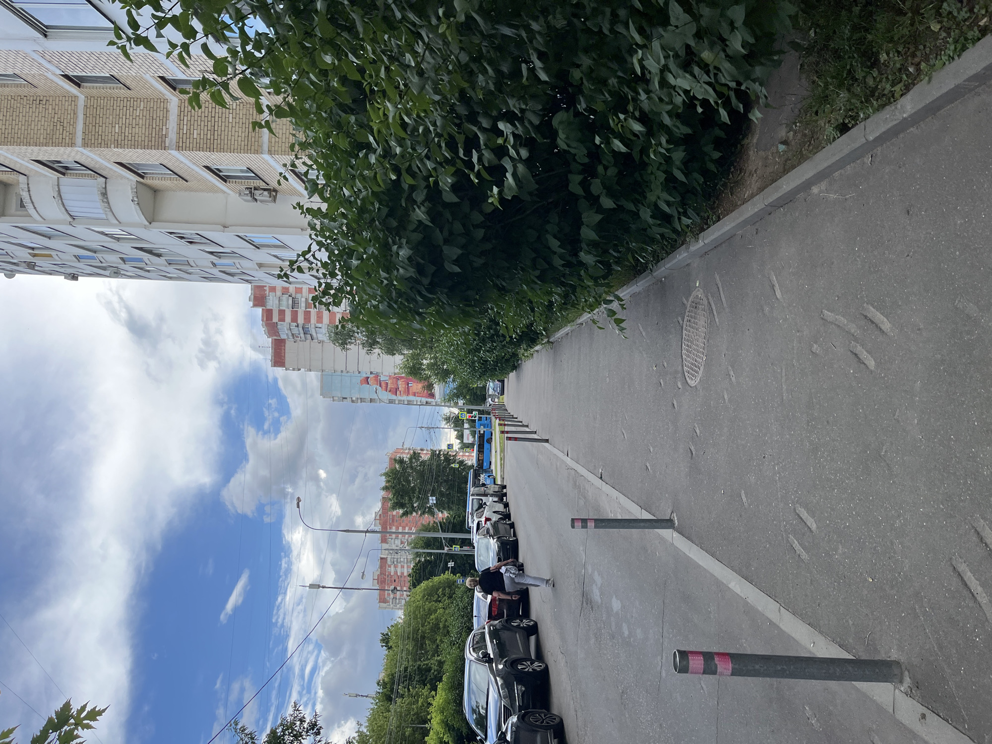 The image depicts a street scene in Moscow, Russia, with a residential building on the right side of the frame. The building is tall and appears to be constructed of brick and concrete. In front of the building is a dense hedge of green foliage. A paved sidewalk runs along the hedge, and further to the left is a street with parked cars. A woman wearing a black t-shirt and grey pants is walking away from the camera on the street. A blue bus is visible in the distance, and a traffic light can be seen. The sky is partly cloudy with a mix of blue sky and white clouds. The overall impression is of a typical urban environment.