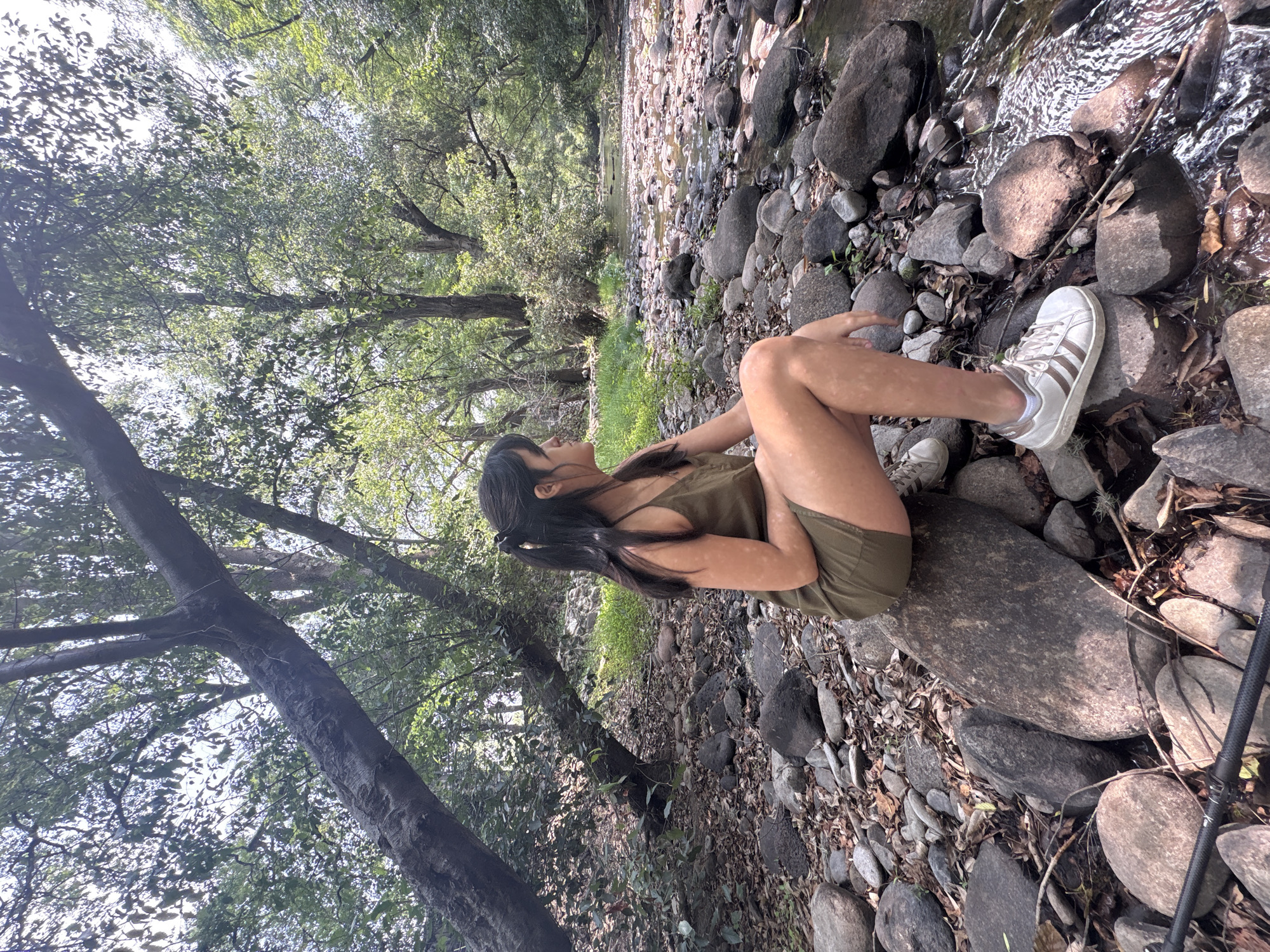 A woman is sitting on a large rock next to a stream in a wooded area in Cañada de Madero, Mexico. The woman is wearing a dark-colored dress and white tennis shoes. She has long, dark hair and is facing away from the camera, looking toward the stream. The stream flows between rocks and trees. The ground is covered with leaves, rocks, and small plants. The trees overhead have green leaves. A fishing rod is located next to the rock in the lower-right portion of the image.