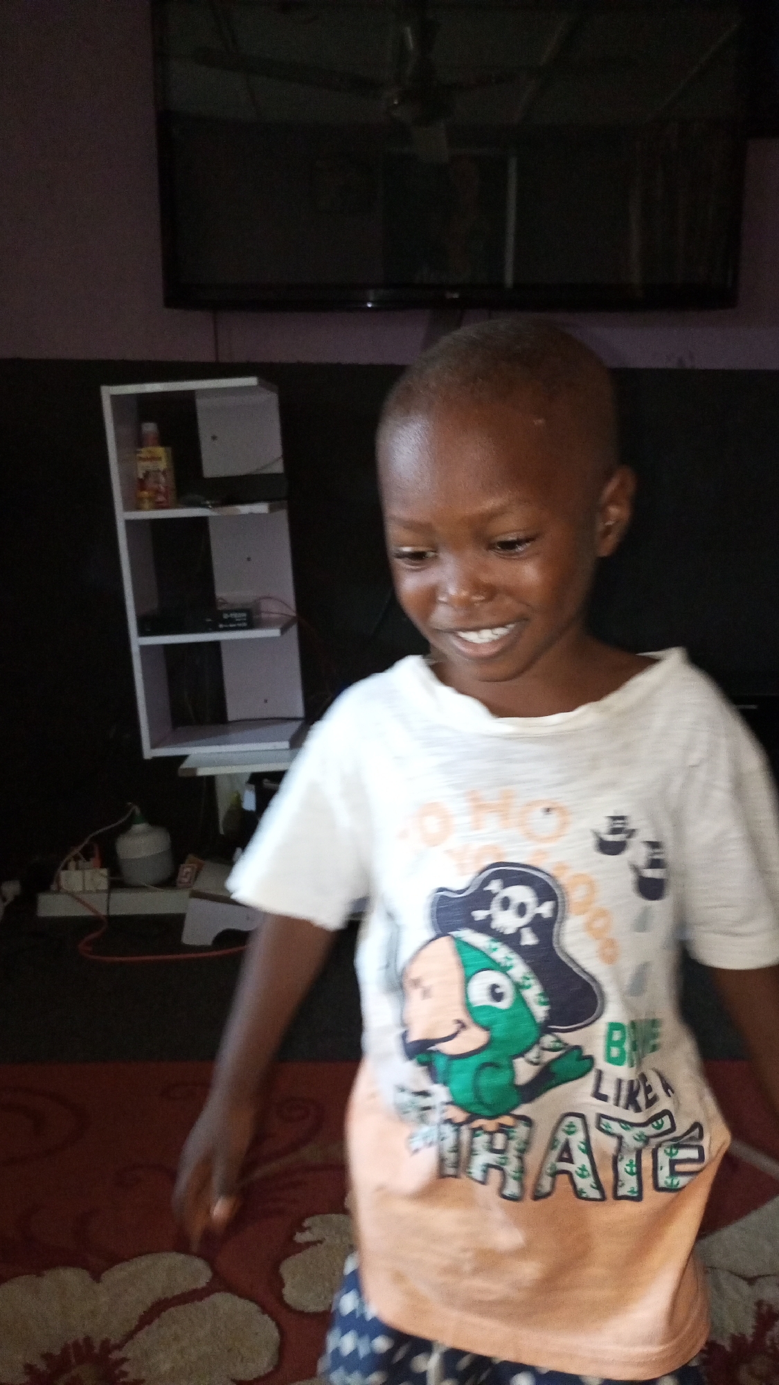 A young child, with dark skin and a short buzz cut, stands in an indoor setting, smiling broadly as they look downwards and to their left. The child is wearing a white t-shirt with a colorful graphic design featuring a pirate skull and crossbones, a green cartoon character wearing a pirate hat, and text that includes "O HO HO" and "PIRATE" (possibly "BORN LIKE A PIRATE" or "BE LIKE A PIRATE"). The bottom part of the t-shirt is a peach/orange color, and they are wearing dark blue patterned shorts with white spots.
The background shows a dark wall with a large, dark-screened television mounted high up. Below the television, to the left of the child, is a white, open-shelved unit. On one of the shelves, a bottle with a red cap is visible, with the word "Paddles" or "Paddlers" partially discernible on its label. Below the shelves, various wires and electrical outlets are visible, suggesting a utility area. The floor is covered by a reddish-brown rug with a light-colored floral or ornate pattern. The lighting appears to be ambient indoor light. The child's face is slightly blurry, indicating movement or a shallow depth of field.