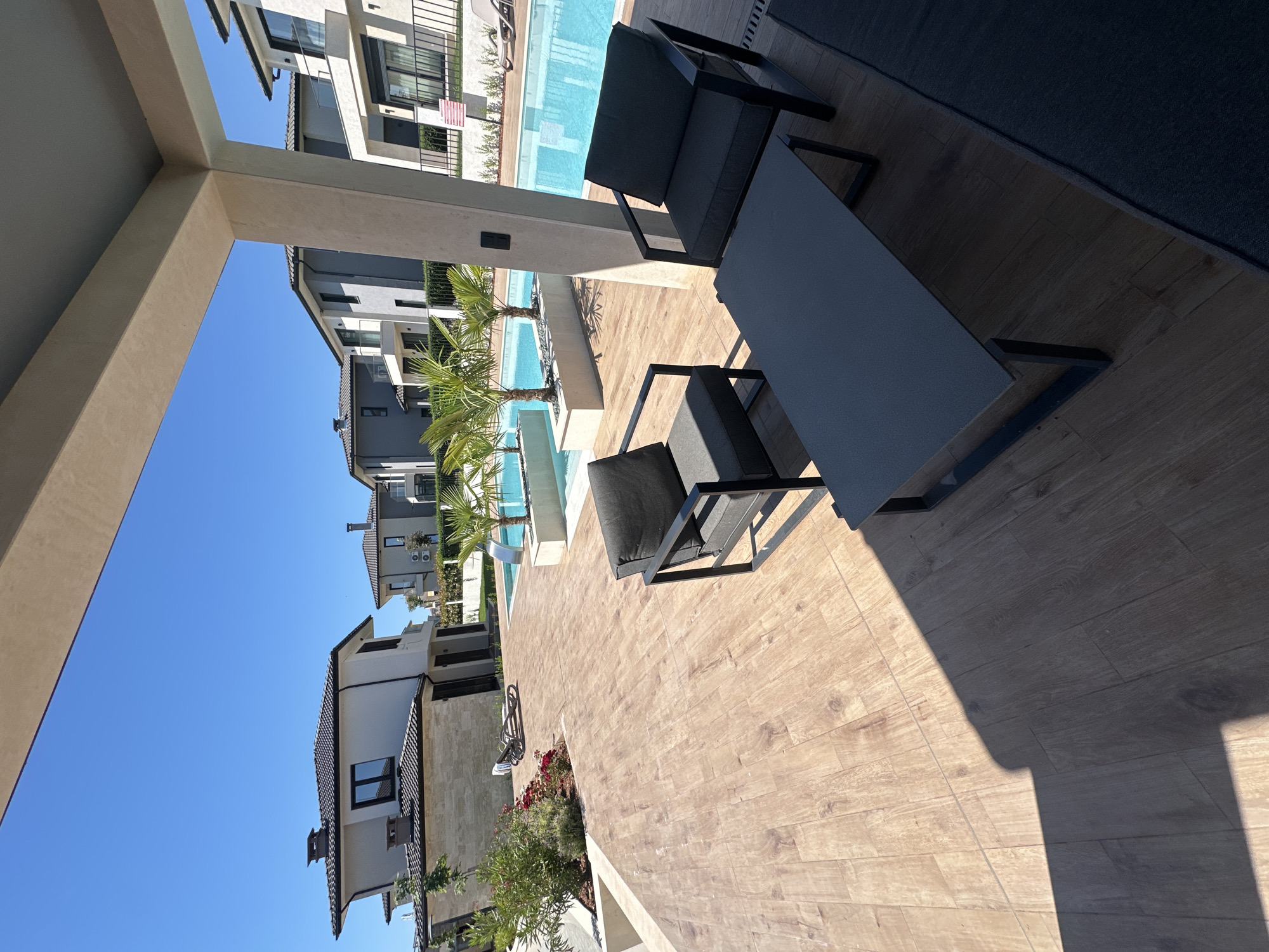 The image depicts a modern residential complex with townhouses and a swimming pool, situated in the city of Pomorie, Bulgaria. The foreground features an outdoor seating area with two dark grey armchairs and a dark grey coffee table, all on a wooden-patterned tiled patio. Behind this area, a bright blue swimming pool is visible, with several palm trees planted in raised beds along its edge. In the background, contemporary townhouses with white and grey facades, dark tiled roofs, and balconies can be seen under a clear blue sky. The scene is bathed in sunlight, casting distinct shadows on the patio. The architecture suggests a newly developed tourist or residential resort.