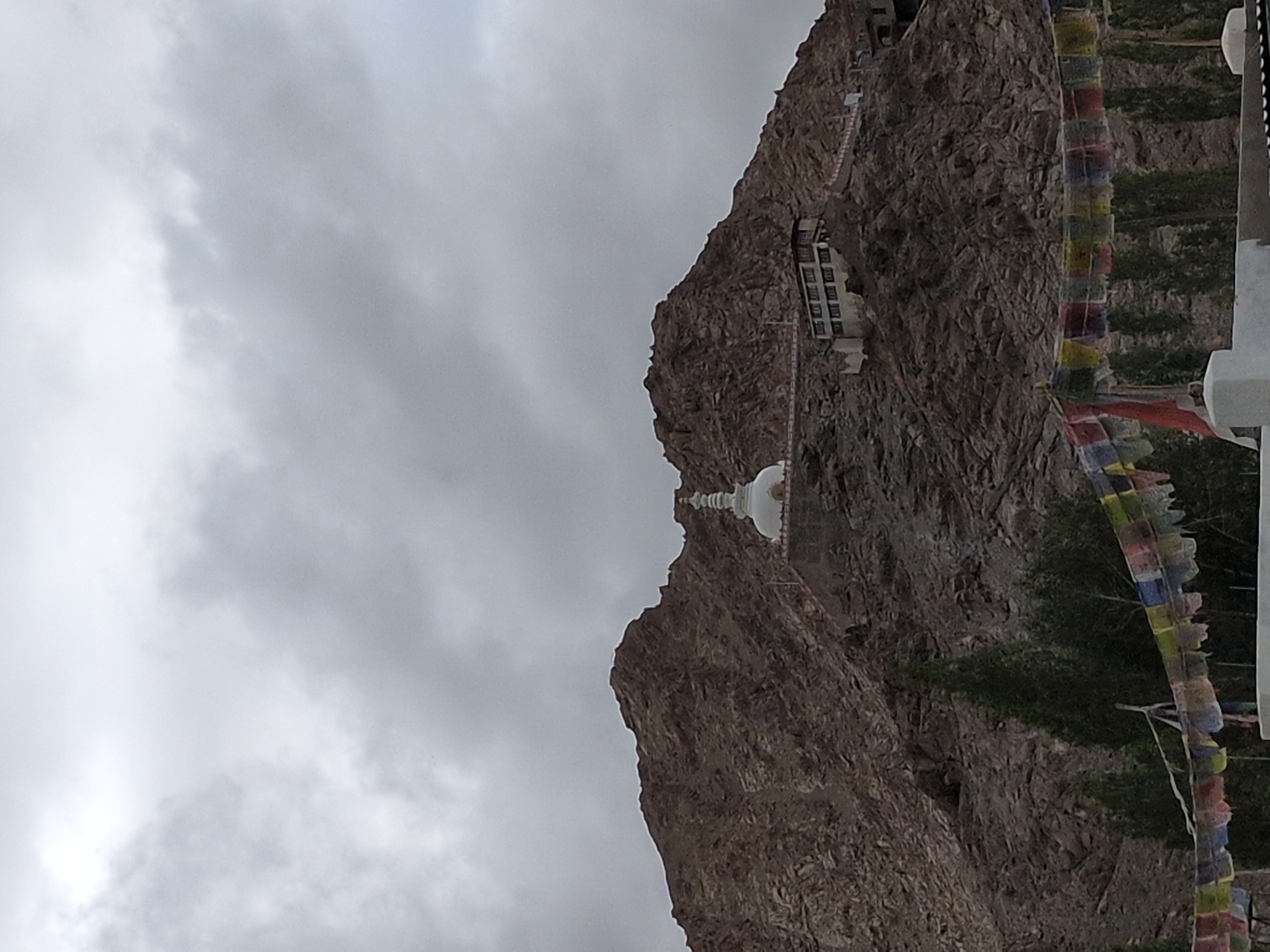 A wide shot captures the Shanti Stupa on a rocky mountain peak in Leh, India, under a cloudy sky. The stupa, a white dome-shaped Buddhist monument, is situated on a raised platform on the left side of the mountain. To the right of the stupa, a multi-story building with white walls and dark windows is built into the mountainside, connected by a pathway. In the foreground, colorful prayer flags are strung across the frame. The mountain itself is rugged and brown, with sparse vegetation and trees dotting the lower slopes and the area in front of the mountain. The overall impression is one of a serene and elevated spiritual site against a natural, imposing backdrop.