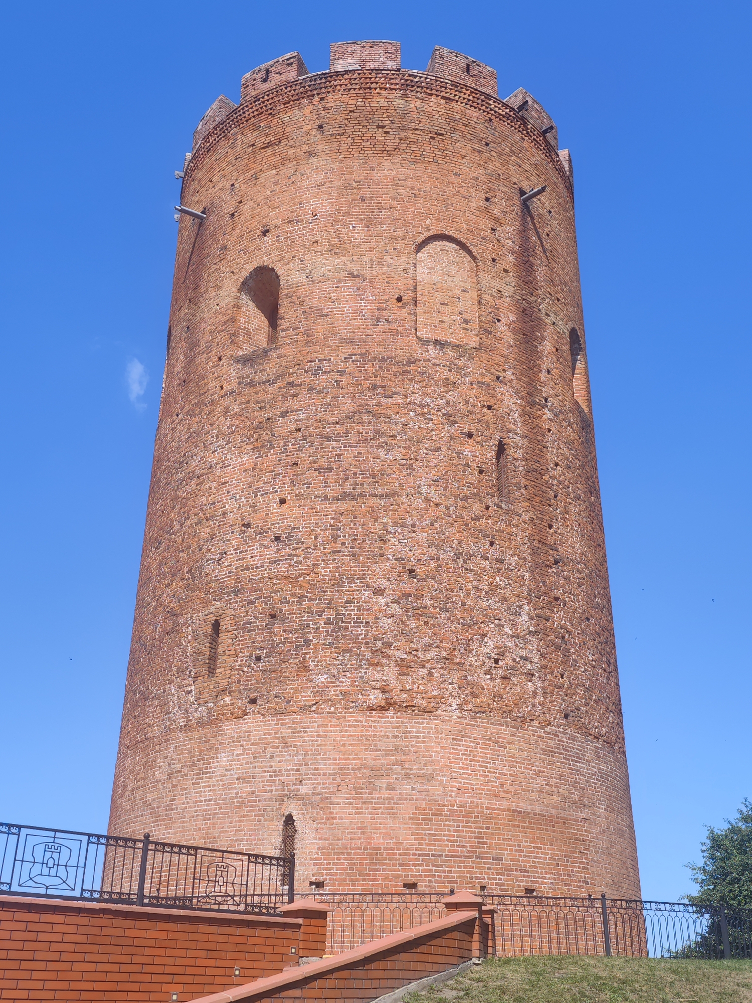 This is a low-angle, full-shot, vertical medium of a cylindrical, brick watchtower against a bright blue sky. The tower is made of reddish-brown bricks, with some areas showing signs of wear and tear. It has a crenelated top and several small, arched openings scattered across its surface. A wooden beam protrudes from the tower near the top, and a metal pipe or conduit runs down the side. At the base of the tower, there is a black metal fence with a decorative pattern, including what appears to be a coat of arms on several panels. The fence is supported by brick pillars and extends across the foreground. Steps made of brick lead up to the fenced area. A small patch of green grass is visible in front of the fence, and a leafy green tree is present on the right side of the frame. The sun is shining brightly, casting shadows and creating a clear, daytime scene. There are no people visible in the image. The overall impression is one of historical architecture in a well-maintained setting. The location is Kamyanyets, Belarus.