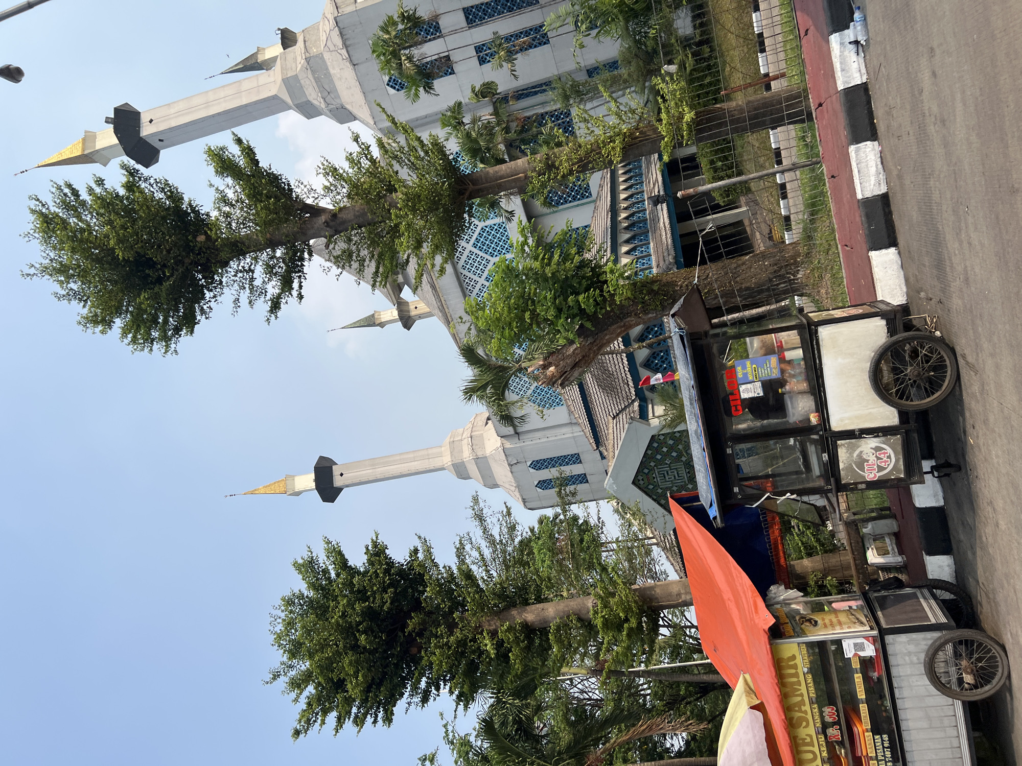 In Gunung Putri, Indonesia, a mosque with tall, white minarets topped with golden spires stands prominently in the background under a clear blue sky. The mosque features intricate blue latticework on its windows. In the foreground, two food carts are parked on the side of a road. The cart on the left has a bright orange tarp covering it and displays a menu with text in Indonesian, including "JUE SAMIR" and prices. The cart on the right, labeled "CIL", is a smaller kiosk with a black and white exterior. Several trees, including tall, slender ones with lush green foliage and palm trees, are scattered throughout the scene, with some growing in front of the mosque and others behind a metal fence. The road in front of the carts has a distinct black, white, and red striped curb. The overall impression is a daytime scene in an urban or semi-urban environment, with a juxtaposition of religious architecture and everyday street commerce.