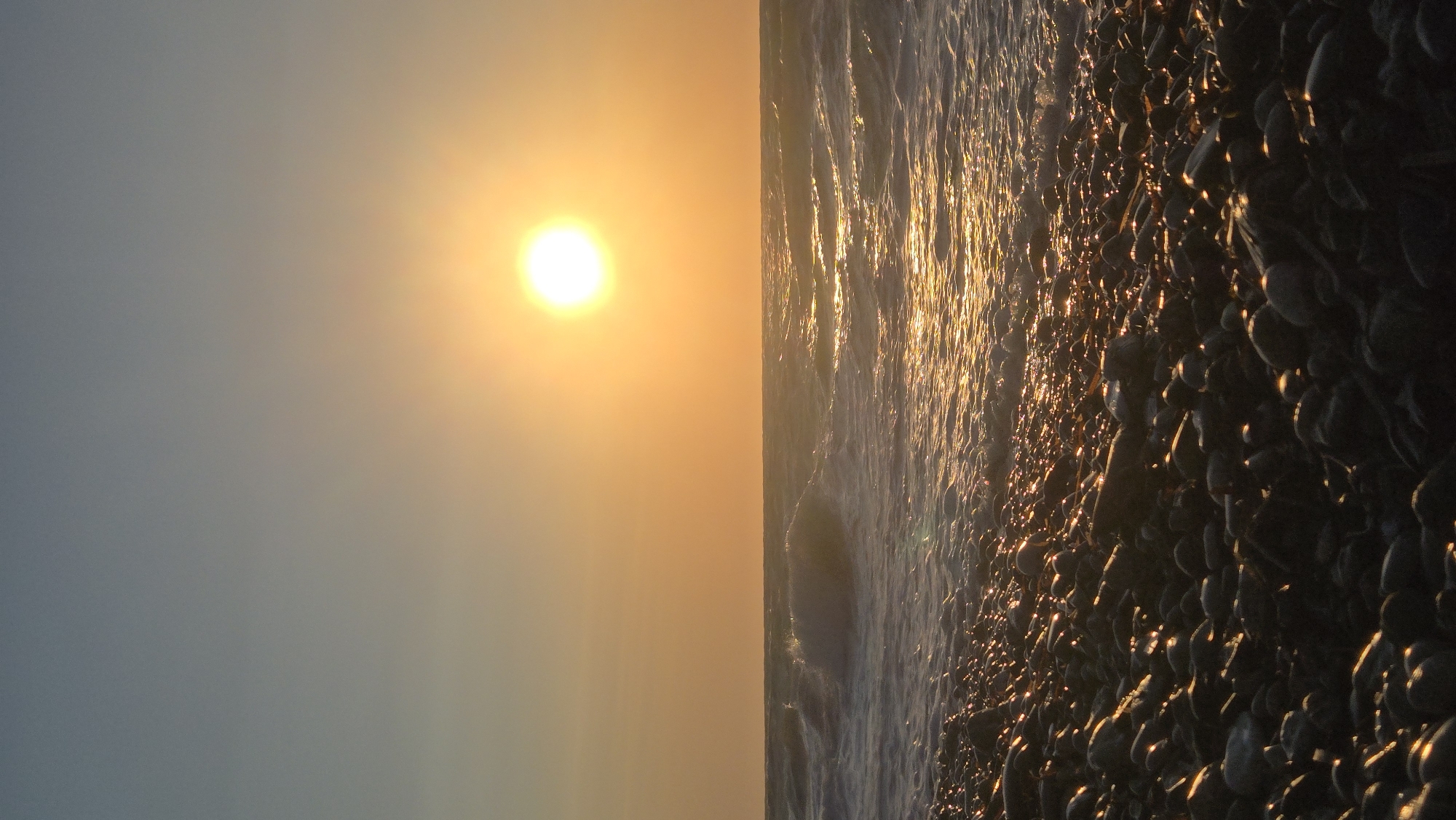 A vertical, wide-angle, medium shot captures a pebble beach meeting the sea under a bright, setting sun. The sky is a gradient of pale blue at the top transitioning to a warm orange near the horizon, where the sun appears as a luminous, white orb slightly to the right of the frame's center. Gentle waves with white crests roll onto the shore, reflecting the sun's golden light. The foreground is densely packed with dark, rounded pebbles, many of which are illuminated by the sun's glare, creating a sparkling effect. No people are visible in the scene, and the environment appears natural and undeveloped. The overall impression is of a serene coastal landscape at dusk.