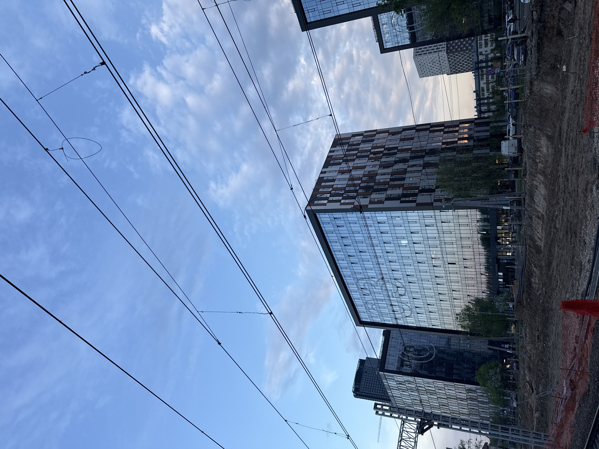 A low-angle shot reveals modern glass-fronted buildings against a backdrop of a blue, cloudy sky. Overhead power lines stretch across the frame, diagonally intersecting with the buildings and the sky. The ground is rough, showing signs of construction or excavation. The location is most likely in Moscow, Russia.