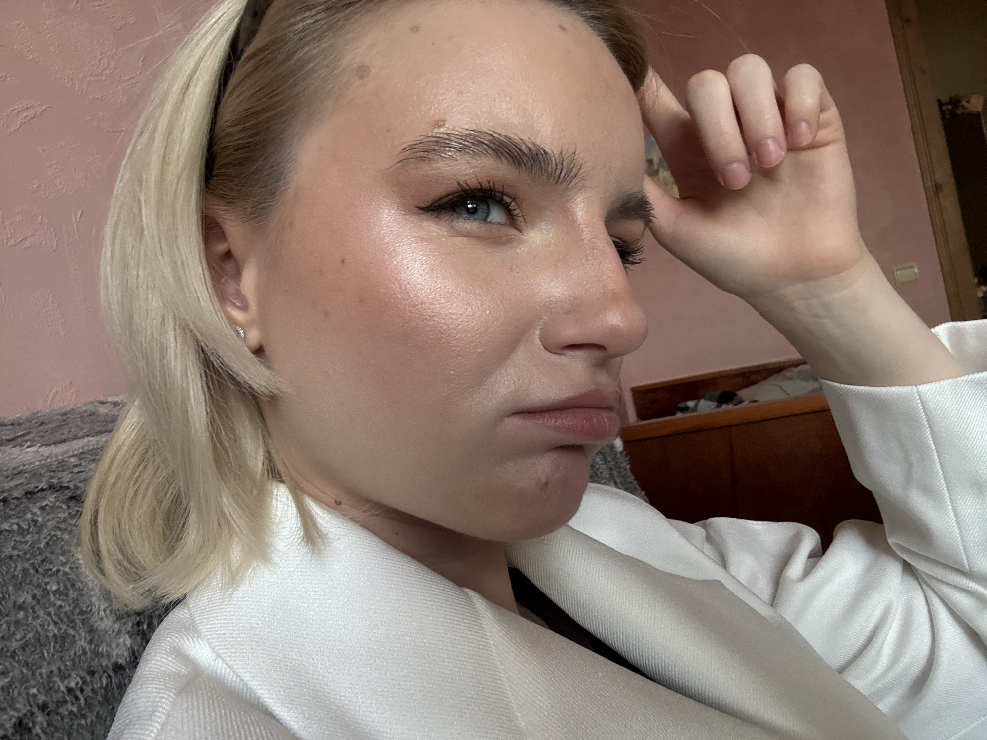 A young woman with blonde hair and blue eyes is captured in a close-up, profile shot, likely indoors in Vsevolozhsk, Russia. She is looking towards the viewer's left with a pensive expression, her lips slightly pursed. Her right hand is raised to her head, with fingers gently curled against her temple, suggesting contemplation or a thoughtful mood.
She has blonde hair, worn with a black headband, and her clear blue eyes are accentuated by dark eyeliner and mascara. Her eyebrows are notably full and well-groomed, appearing laminated. Her skin has a dewy finish, with blush and highlighter visible on her cheekbones. A few small moles are present on her forehead and neck, and a minor blemish is visible on her chin. She is wearing a white, textured jacket or blazer. A small, stud-like earring is visible in her left ear, and her fingernails are natural and unpolished.
The background features a textured, light pink wall on the left and a portion of a dark grey, plush fabric, possibly a blanket or cushion, in the lower left. Towards the right, dark wooden furniture, likely a cabinet or dresser, is partially visible, along with a light-colored rectangular object on the wall. The lighting is soft and even, indicating an indoor setting.