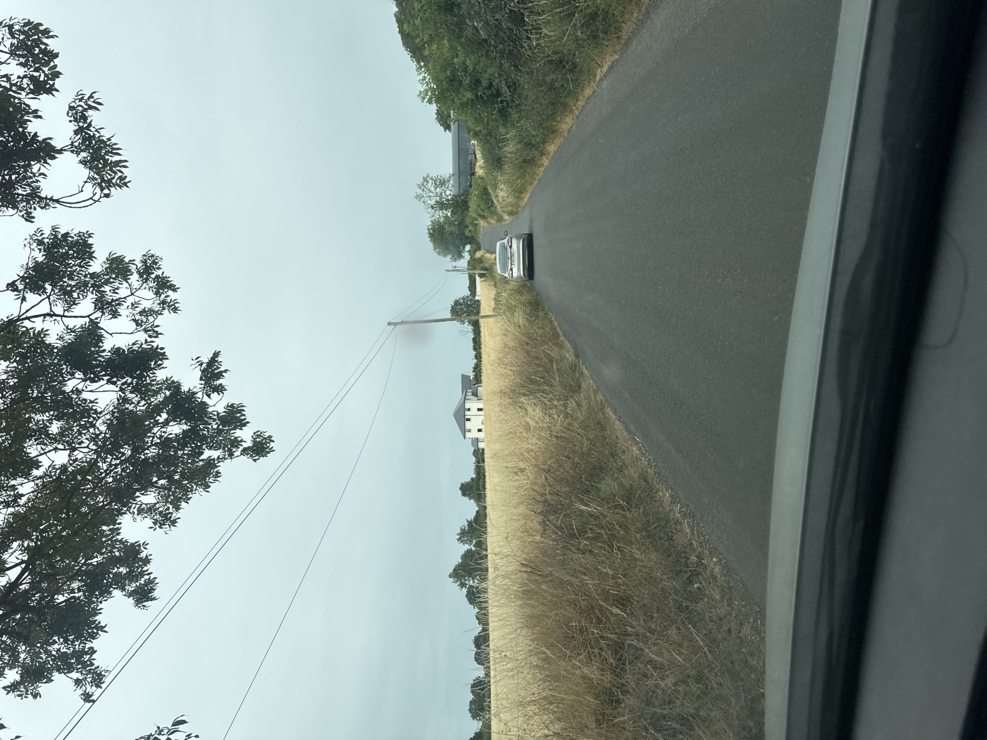 An overcast day in rural Ireland shows a silver sedan driving down a narrow asphalt road. The road is flanked by tall, dry grass on the left and lush green vegetation on the right. In the distance, a white house with a dark roof sits behind a field of golden wheat. Power lines stretch across the sky, supported by wooden poles. Trees line the horizon, and a large, dark structure, possibly a barn or shed, is visible behind the greenery on the right. The photo appears to be taken from the perspective of a vehicle, with the dashboard visible in the foreground.