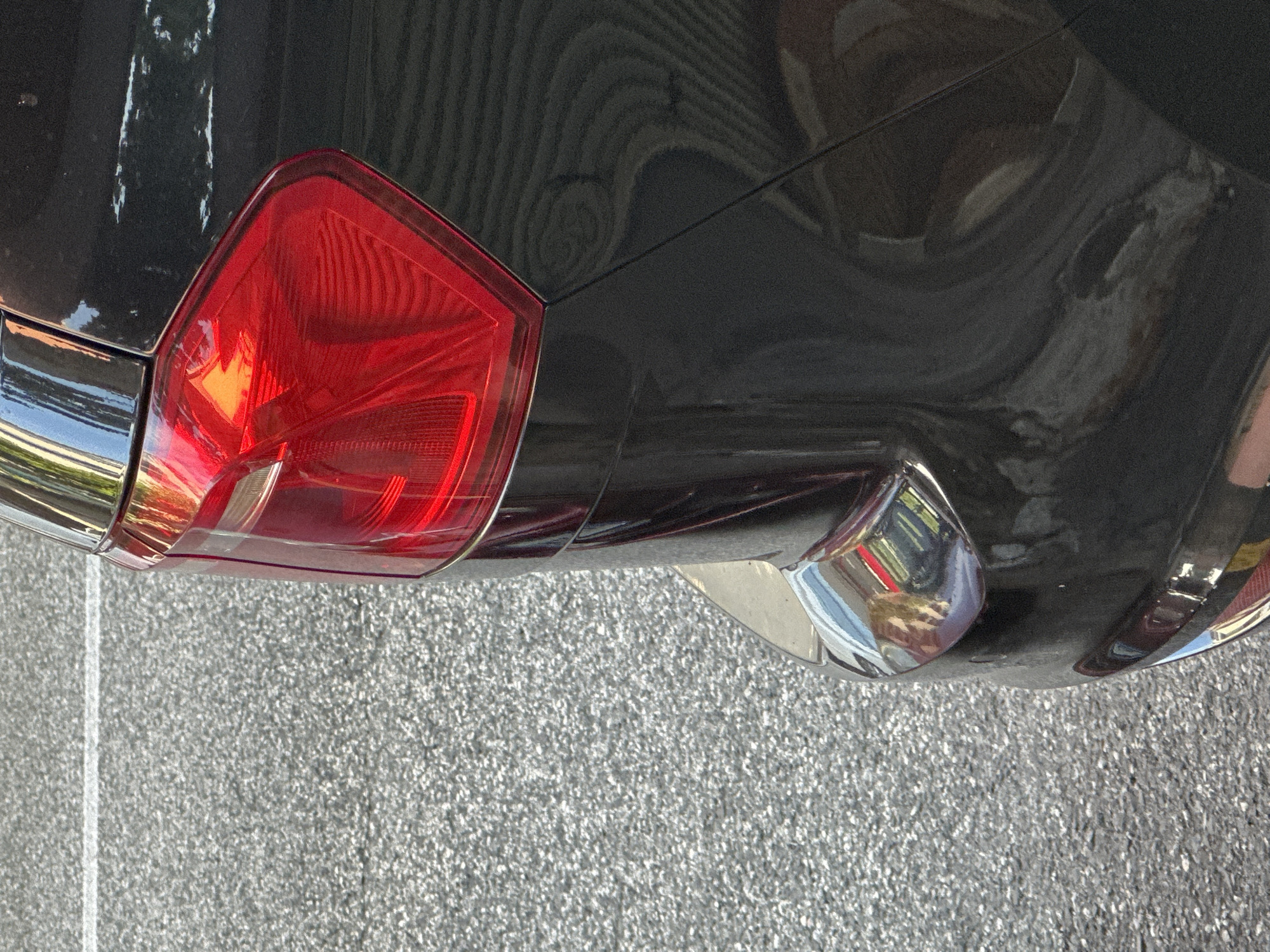 The image is a close-up, exterior shot of the rear portion of a dark-colored vehicle, likely a car, parked on asphalt with a white line visible in the upper left quadrant. The vehicle's red taillight is prominently displayed, featuring a faceted design with internal reflective elements. A portion of the car's bumper, adorned with chrome trim and reflecting the surrounding environment, is visible in the lower part of the frame. The background consists of textured asphalt, characteristic of a street or parking area. The scene is illuminated by natural light, suggesting an outdoor setting, possibly in Trieste, Italy, given the context. There are no people or discernible actions depicted in the image.