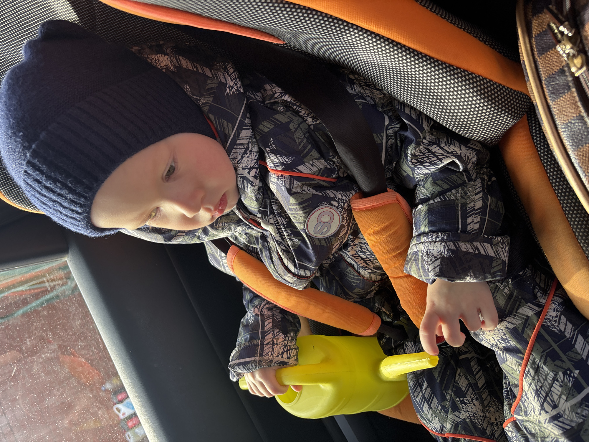 Inside a vehicle in the city of Ферма, Russia, a young child is seated in an orange and black car safety seat. The child, wearing a navy blue knit beanie and a patterned dark blue and green winter suit with red piping, holds a bright yellow plastic watering can in their left hand. Orange padded covers are visible on the car seat's shoulder straps, which secure the child with a black harness. The child is looking downward and to their right. Through the car window behind the child, a blurry reddish-brown exterior is visible, with faint outlines of what appear to be plastic bottles along the lower edge. A brown patterned object, possibly a bag, is partially visible in the lower right foreground. Natural light illuminates the scene from the window side.